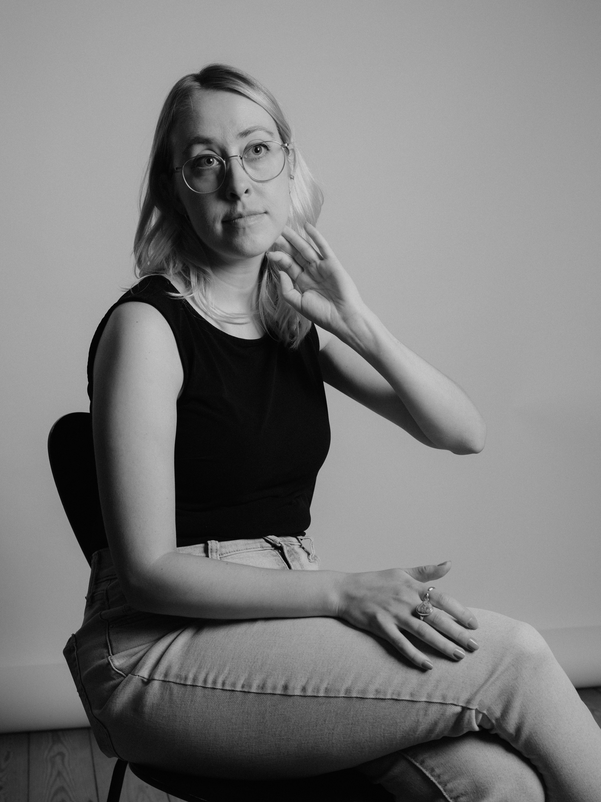A woman in glasses and a sleeveless top sitting on a chair, with crossed legs, with her right hand near her neck and her left hand resting on her knee, in a black-and-white photograph.