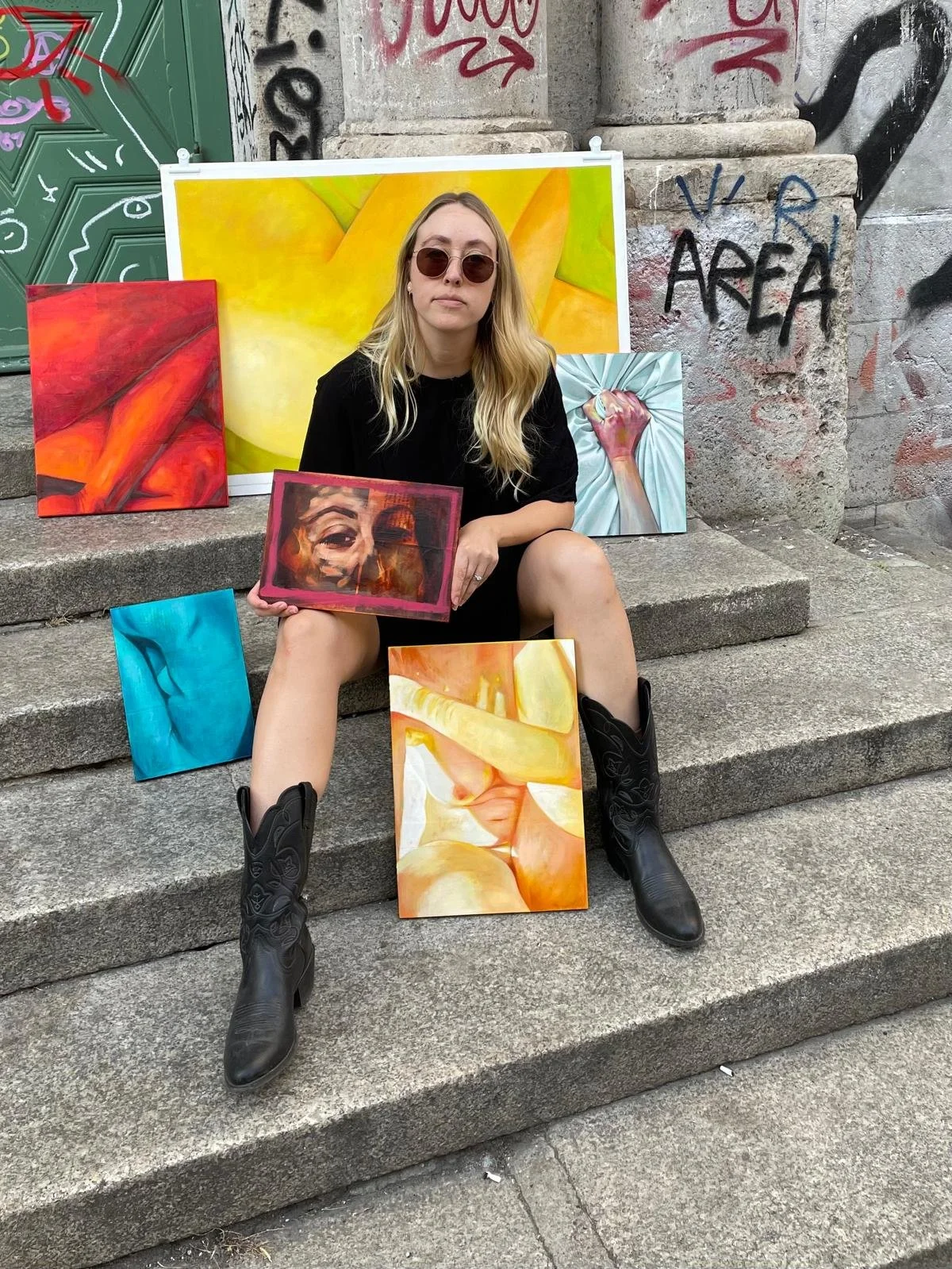 A woman wearing sunglasses and cowboy boots sitting on steps outdoors, surrounded by colorful paintings and artworks displayed behind her, with graffiti on the wall.