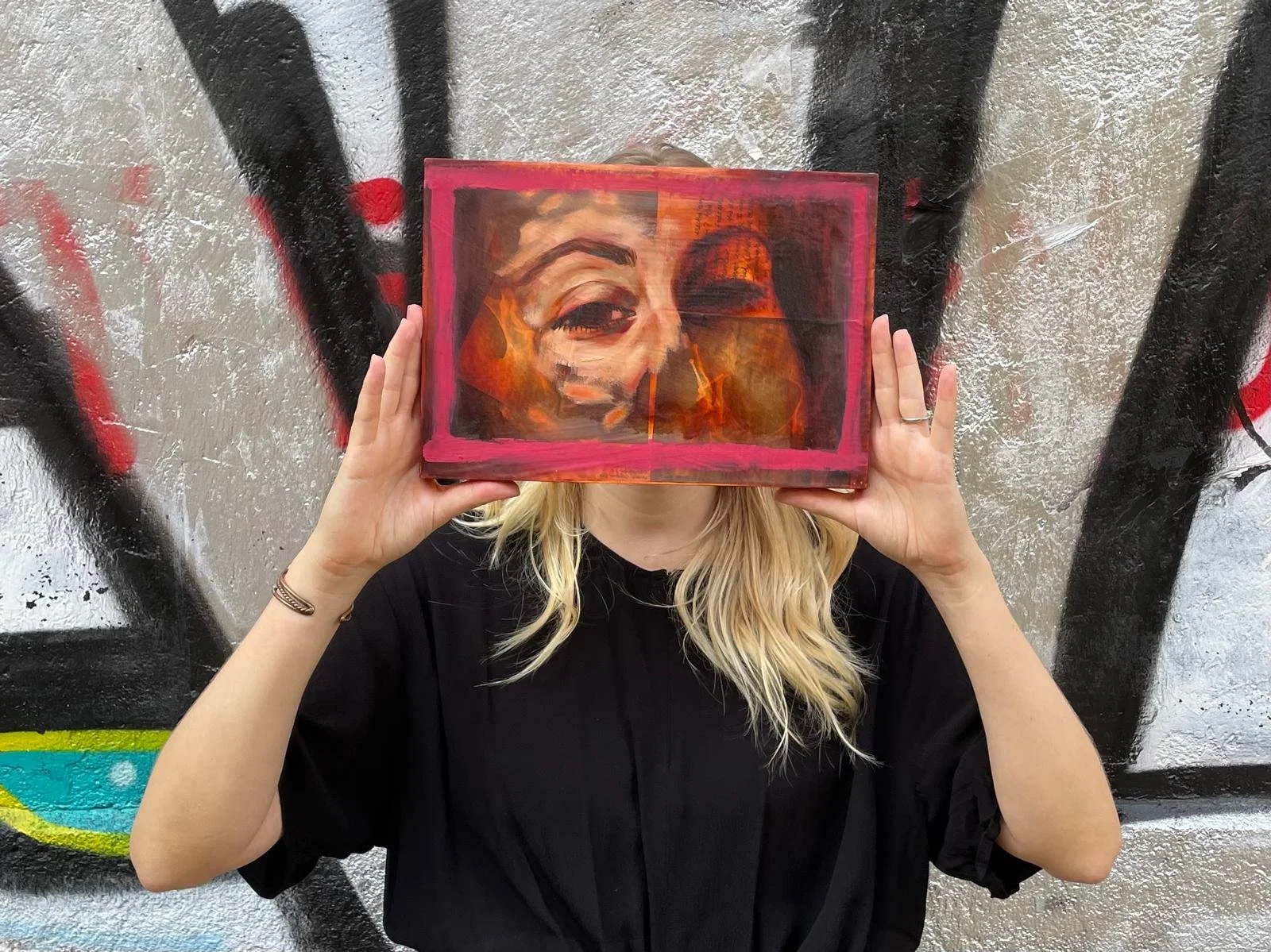 A person with blonde hair holding a painting of a woman's face framed in pink in front of her face. The background is a graffiti-covered wall.