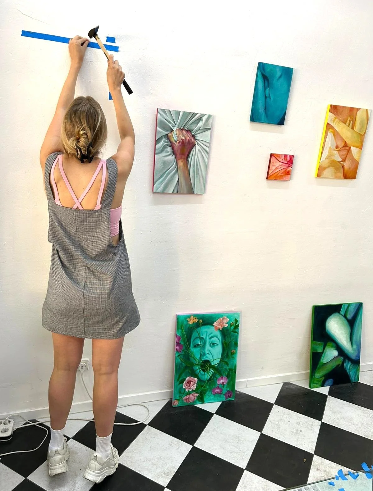 A woman hanging paintings in an art gallery with a black and white checkered floor. She is holding a hammer and thumbtacks, fixing a painting on the wall. The wall features several colorful artworks, including a woman's face surrounded by flowers, a clenched fist on fabric, and other vibrant paintings.