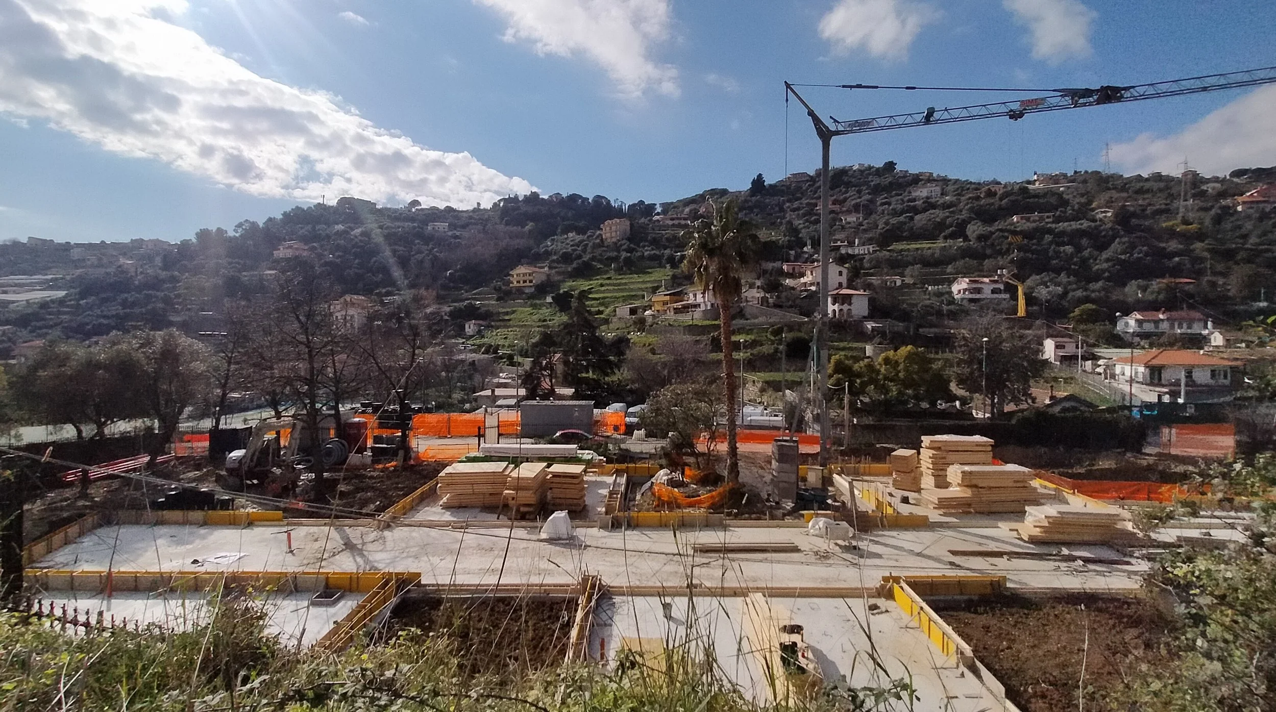 Construction site with foundation work in progress, stacked wood, construction materials, a crane, and hillside houses in the background under a partly cloudy sky.
