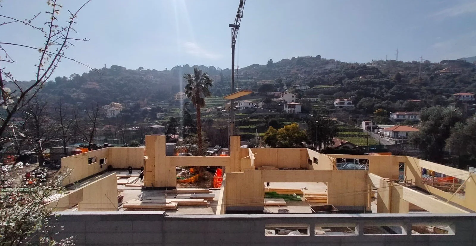 Construction site with wooden framing for a house, surrounded by a bunch of trees and overlooking a hillside neighborhood with houses and a crane in the background.
