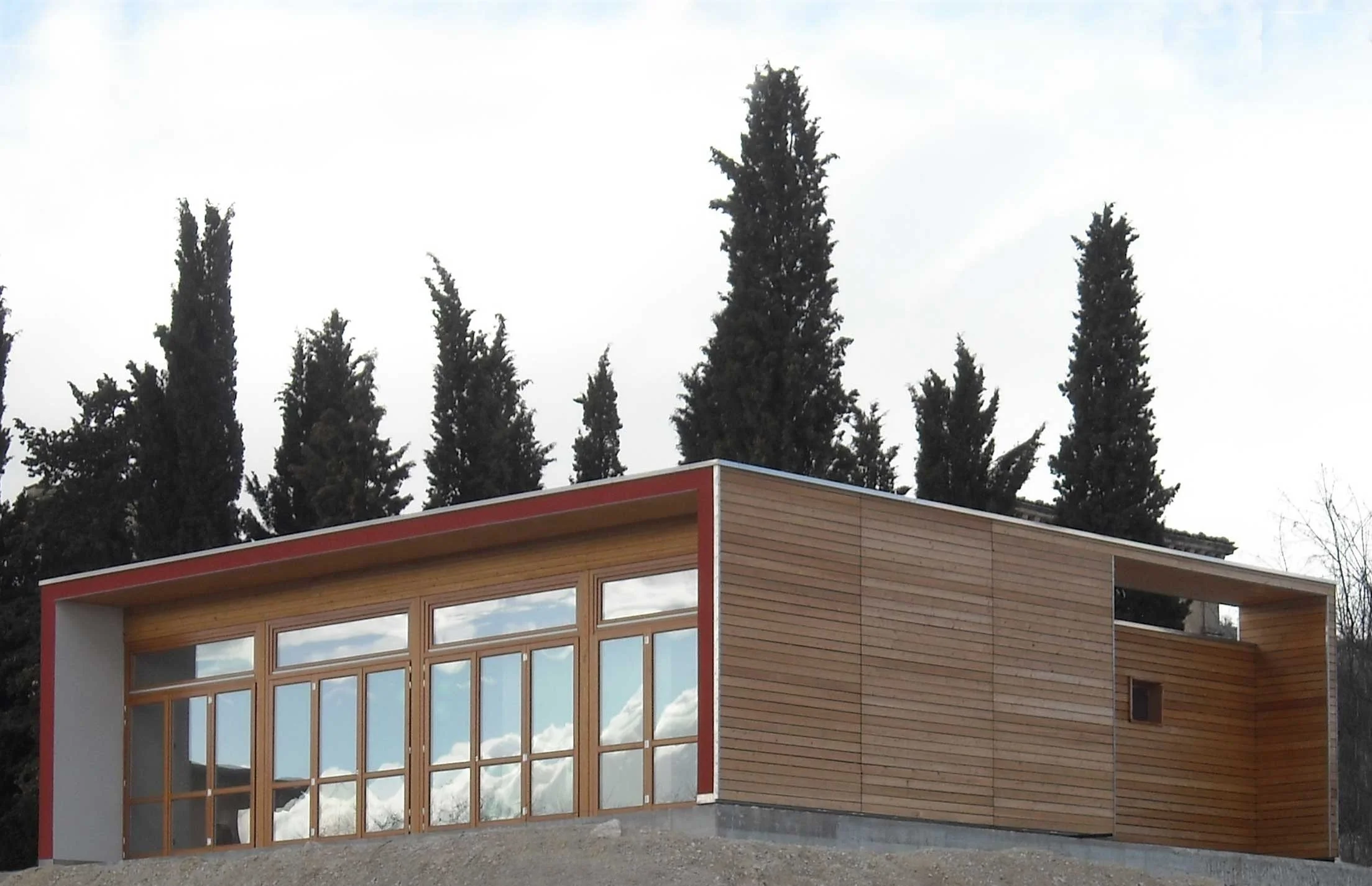 Modern building with large glass windows, wooden paneling, and red trim at the eaves, set against tall trees and a partly cloudy sky.