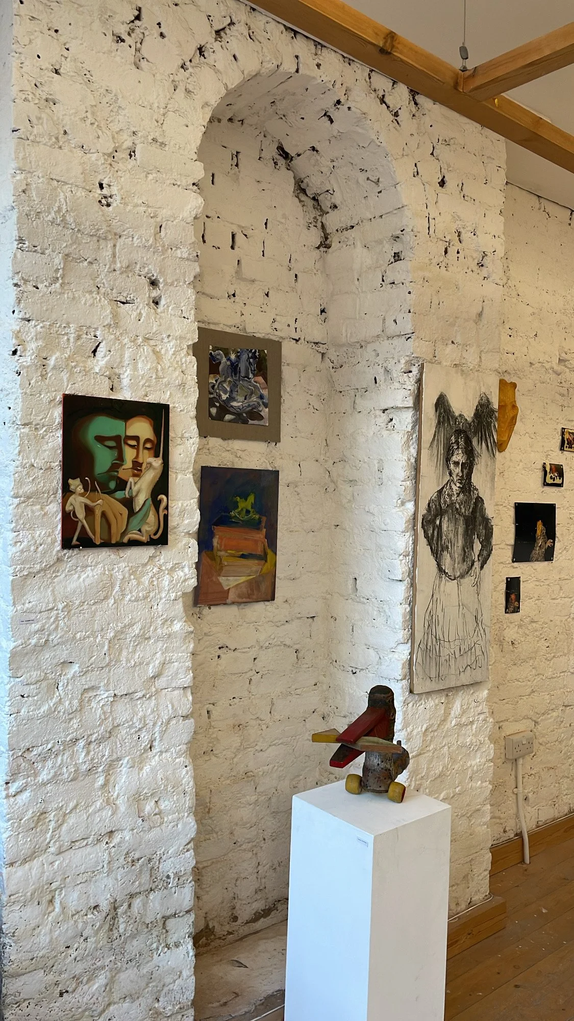 Art gallery with white brick walls displaying various paintings, including abstract and portrait art, with a wooden sculpture on a white pedestal in the foreground.