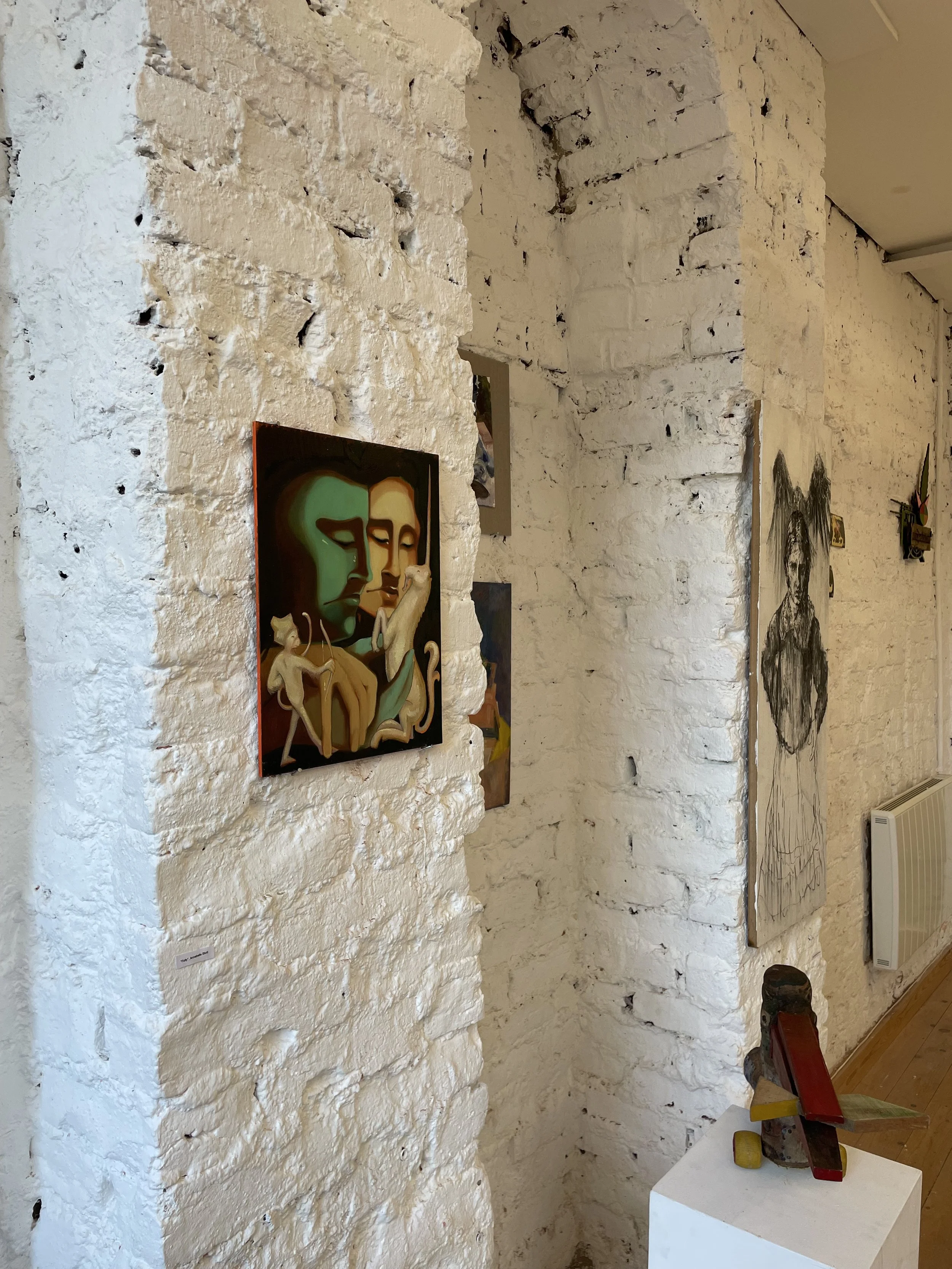 Art gallery with exposed white brick walls displaying colorful paintings, including stylized faces. Artworks are mounted on the wall, and a small white pedestal with sculptural objects and a wooden plank is in the foreground. A radiator is visible at