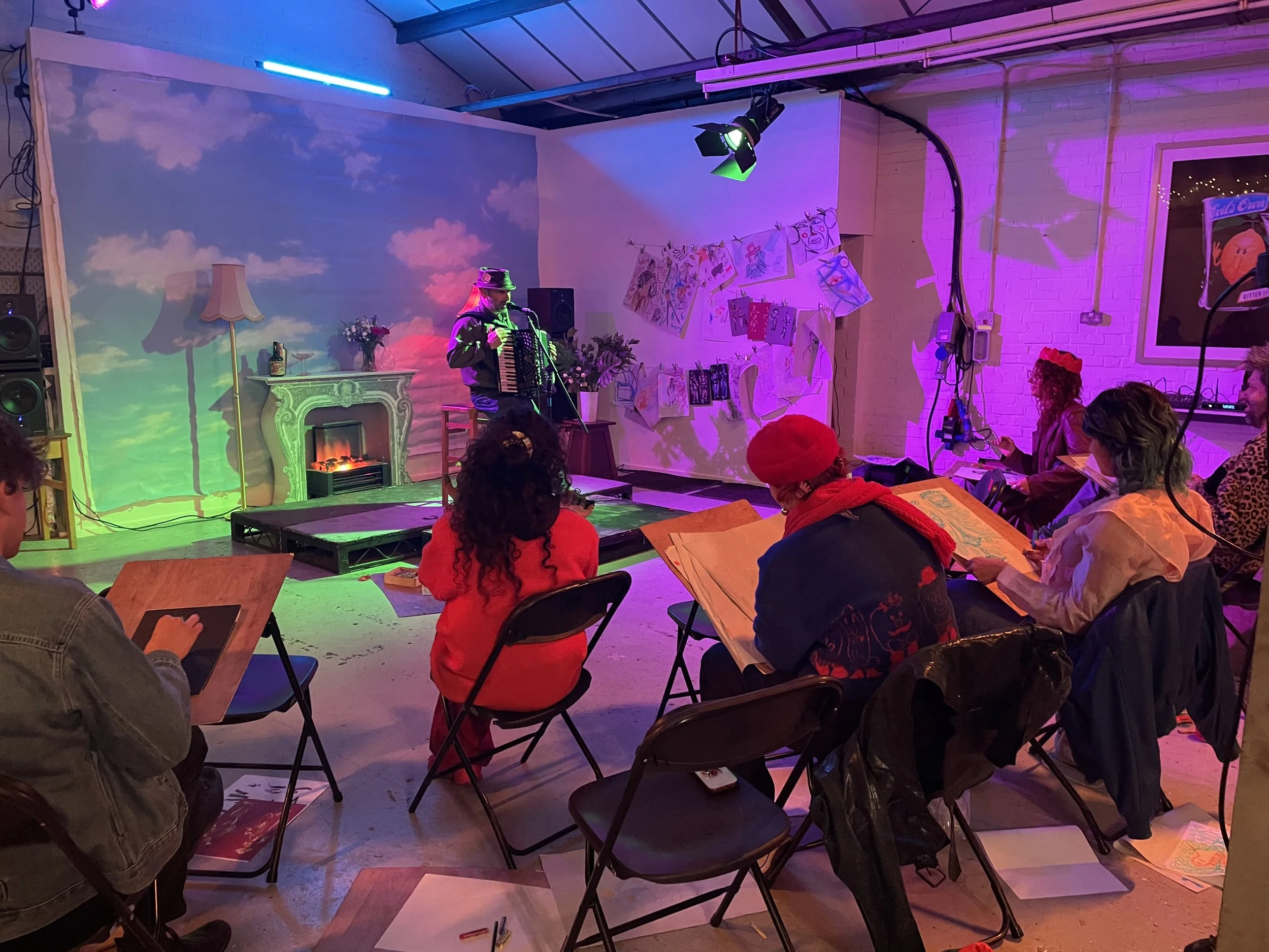 An indoor event with a performer playing an accordion on a small stage, surrounded by colorful lighting and an audience seated and reading papers.