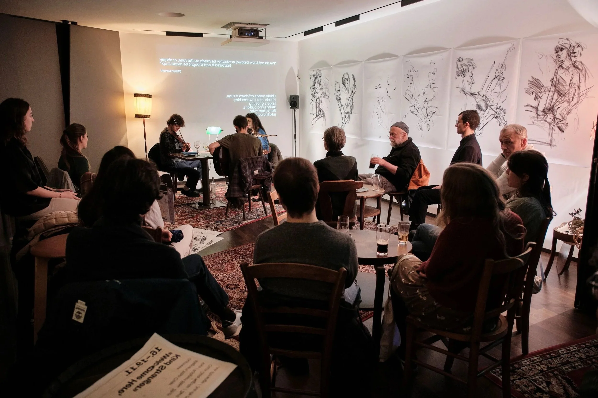 People attending a live music performance in a cozy, dimly-lit venue. Musicians playing violins on stage, with artwork on the wall and an audience seated and watching.
