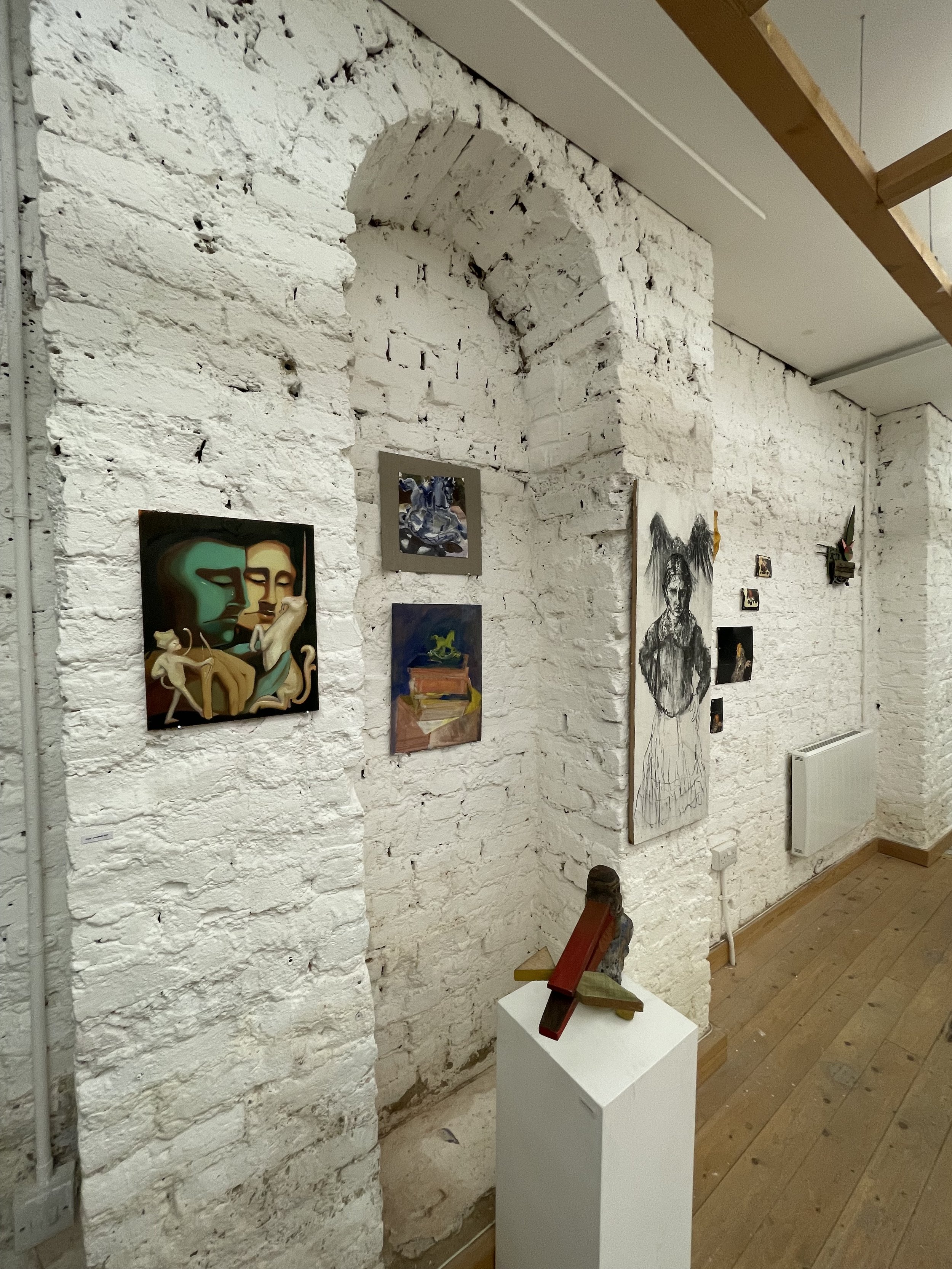 Art gallery with white brick walls displaying paintings. In the foreground, a white pedestal supports wooden sculptures, with wooden flooring and ceiling beams visible.