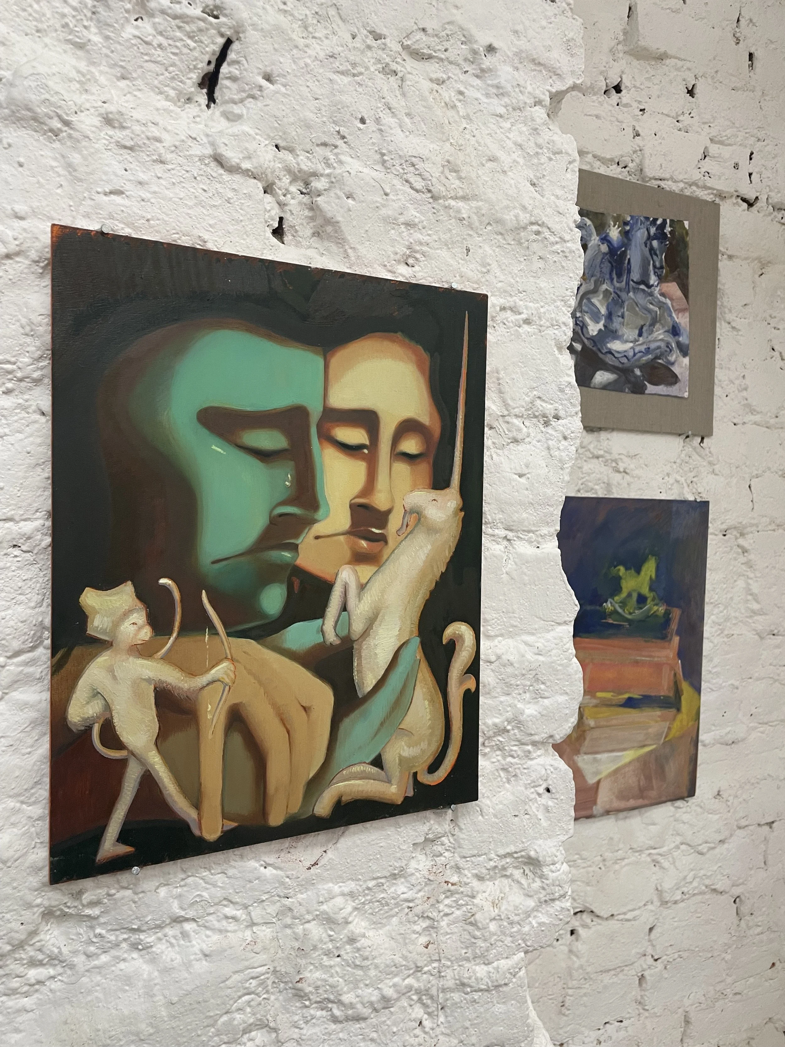Paintings displayed on a white brick wall in an art gallery, including a portrait of a woman with closed eyes and other abstract artworks.
