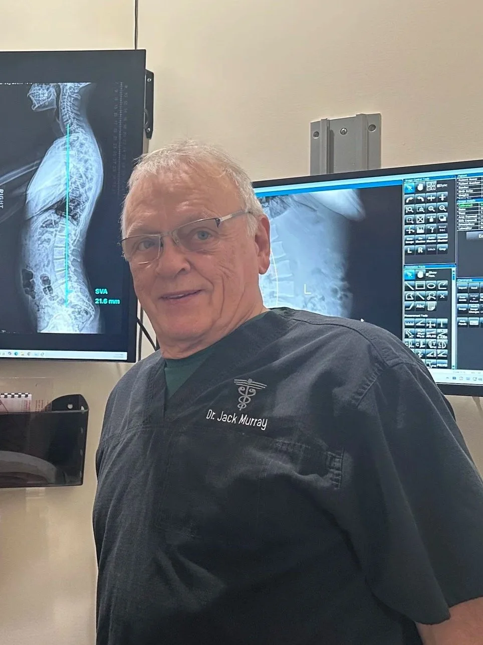 Dr Murray with medical monitors displaying spinal X-rays in the background explaining chiropractic care to a sciatica patient.
