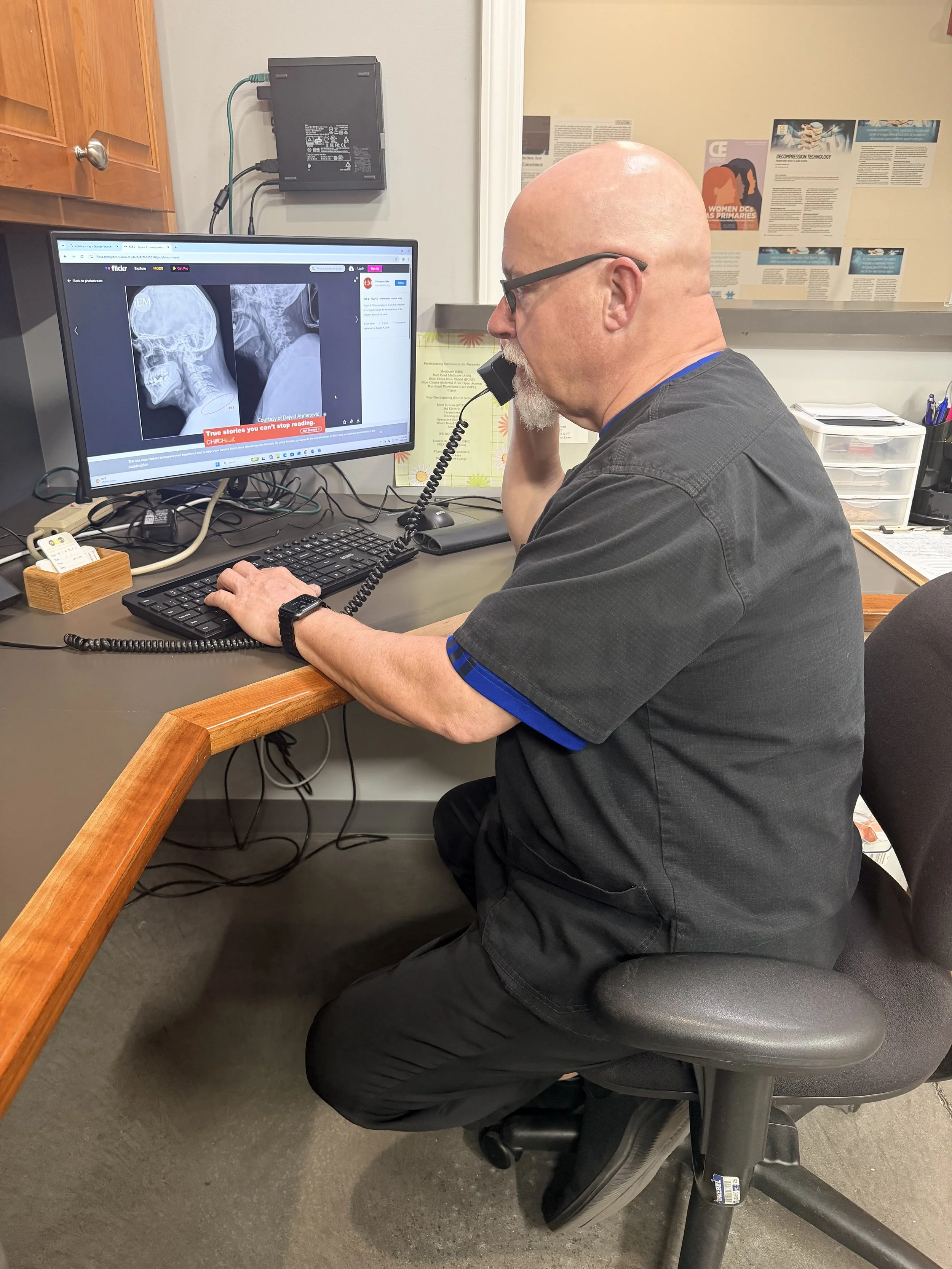 Dr Bohn sitting at a desk, talking with a patient for a free consult about their neck pain, looking at a monitor displaying X-ray images of a skull and neck.