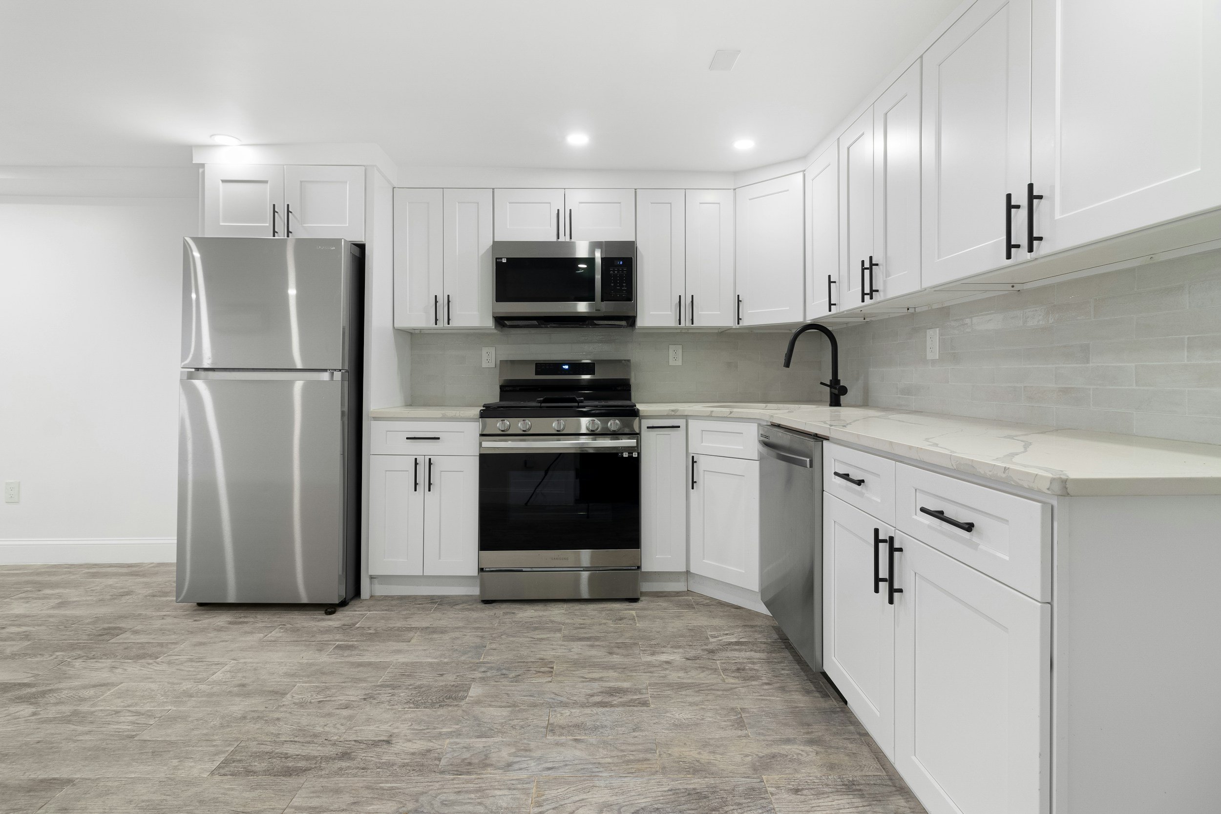 Modern kitchen with white cabinets, stainless steel refrigerator and microwave, black handles, gray backsplash, marble countertop, black faucet, and gray wood flooring.