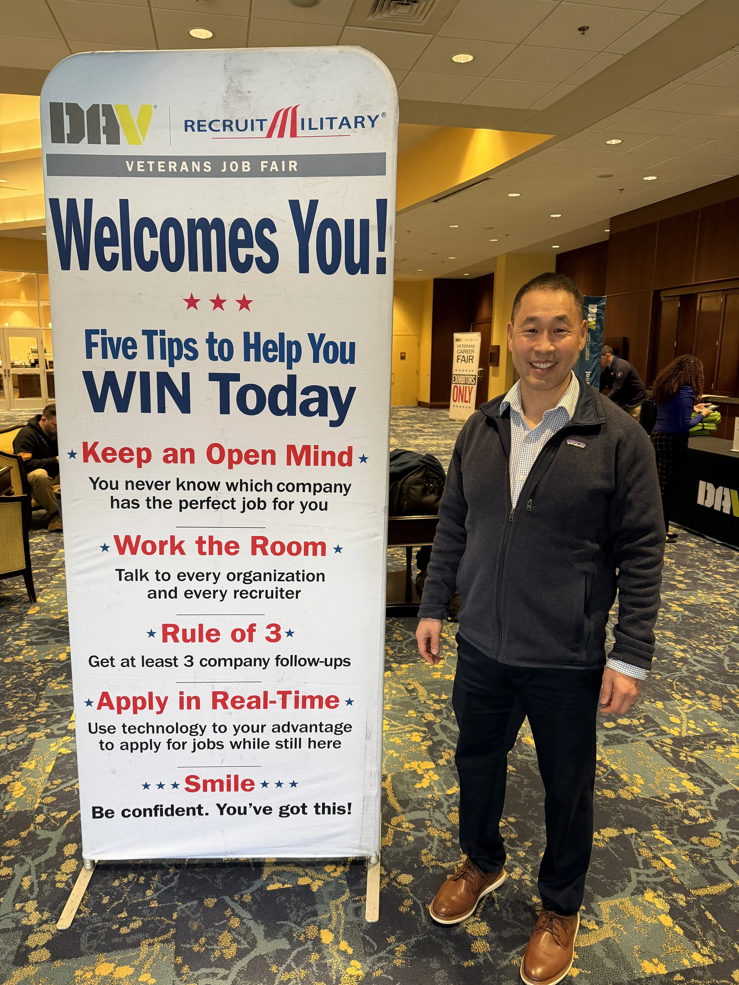 A man standing beside a tall veterans job fair sign in an indoor conference center. The sign displays tips for job seekers, with a man dressed in a collared shirt, dark pants, a dark jacket, and brown shoes smiling at the camera.