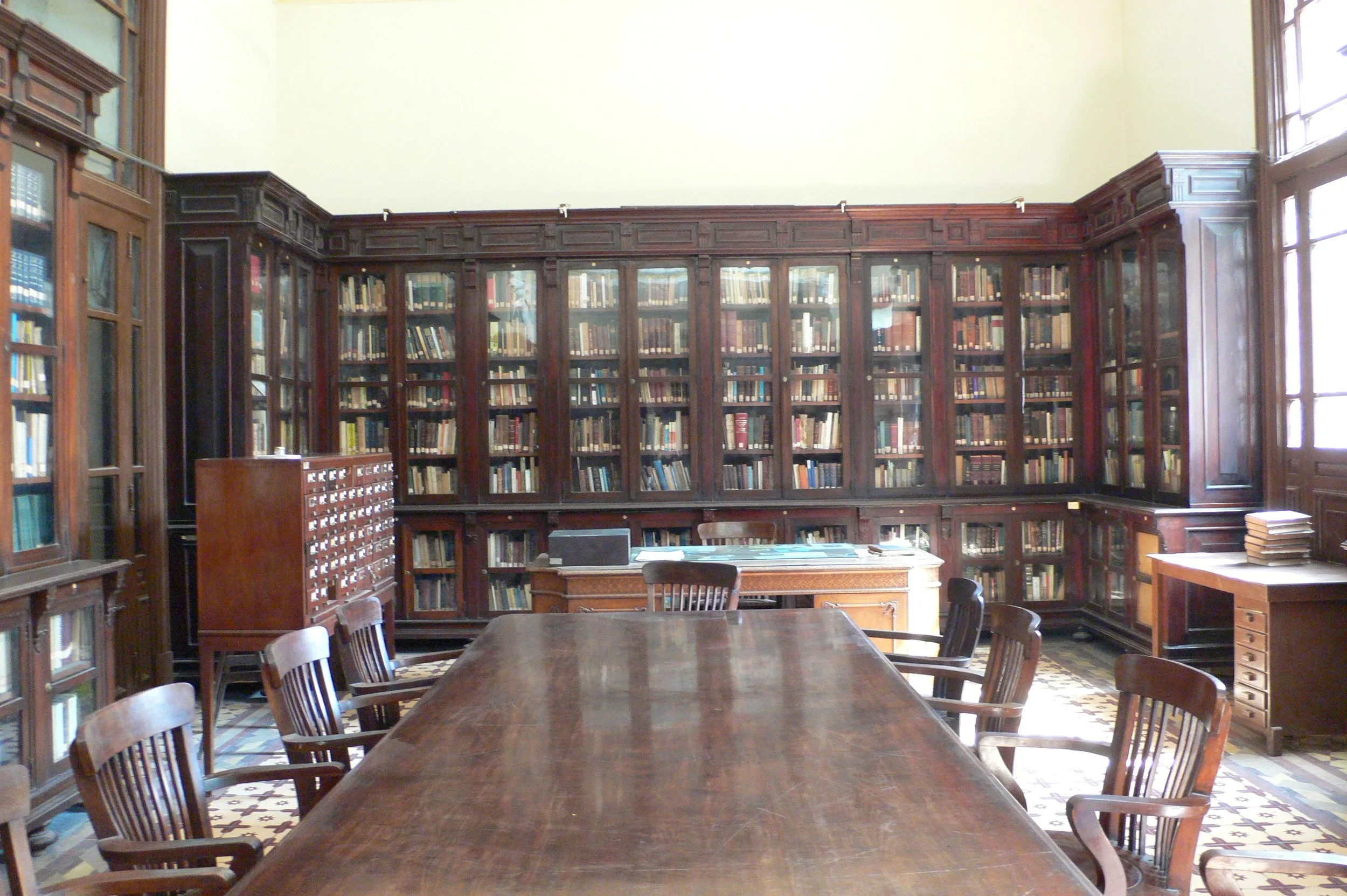 Tall, dark-wood bookshelves with glass doors span the wall. A card catalog sits in front and to the left of the bookshelves; a glass-covered desk appears to the right.