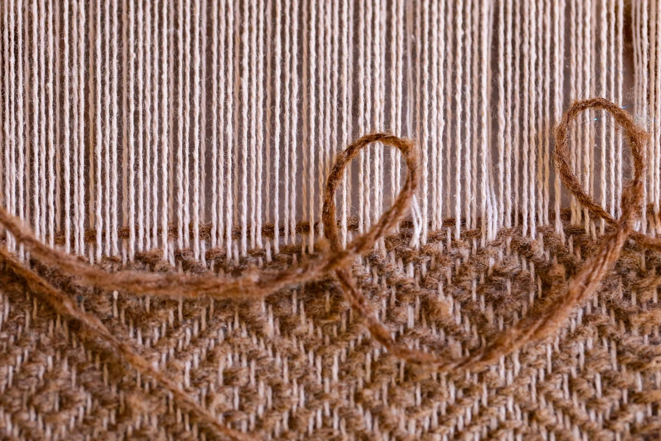 A close-up photo of yarn on a wool string loom. The warp is a light-colored yarn, while the weft is a light brown. The weaving is only partially complete.