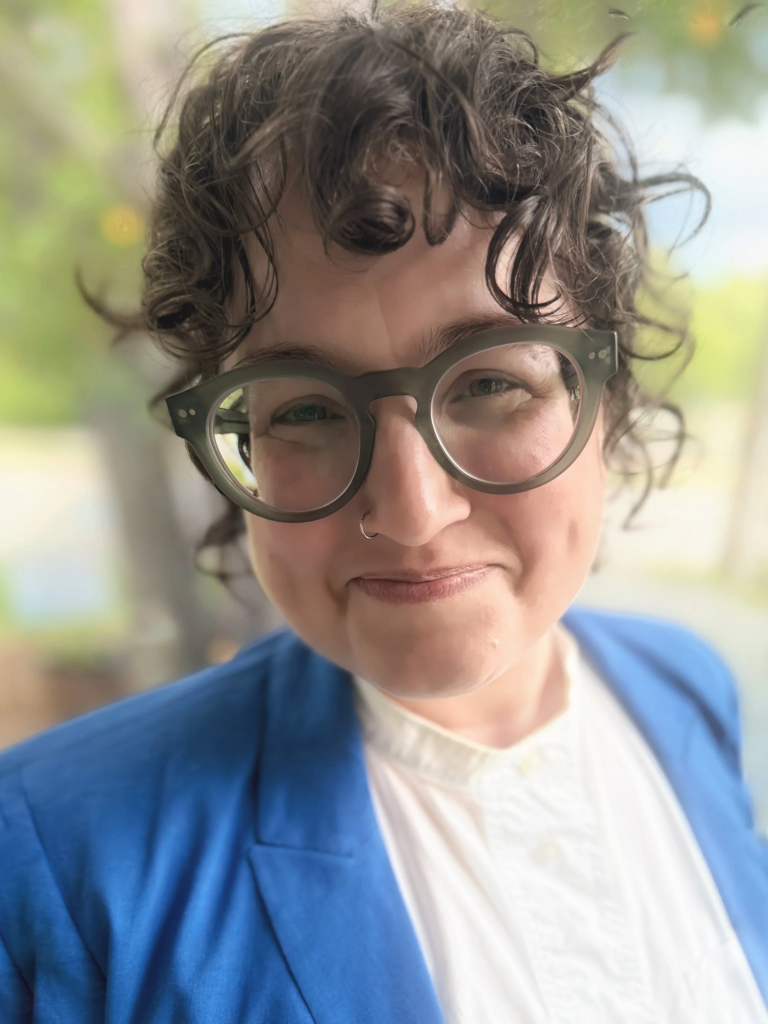 Close-up of a white person with curly hair pulled back and green glasses. She is wearing a white shirt and blue suit jacket and smiling.