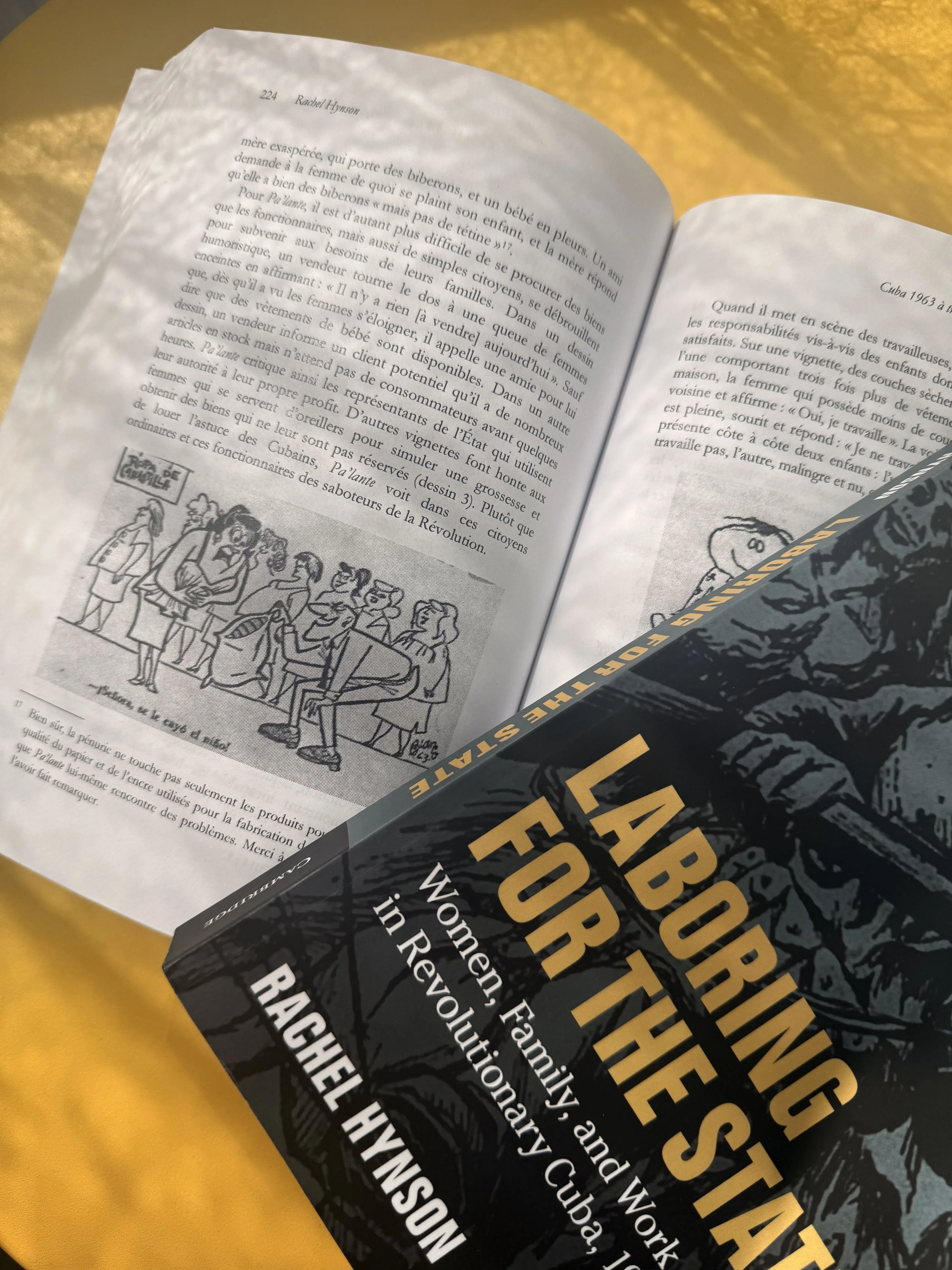 Journal issue is spread open on a yellow surface with text in French and a drawing of a gathering of people. Partially visible too is the cover of a book, Laboring for the State by Rachel Hynson, featuring the title in bold yellow letters.