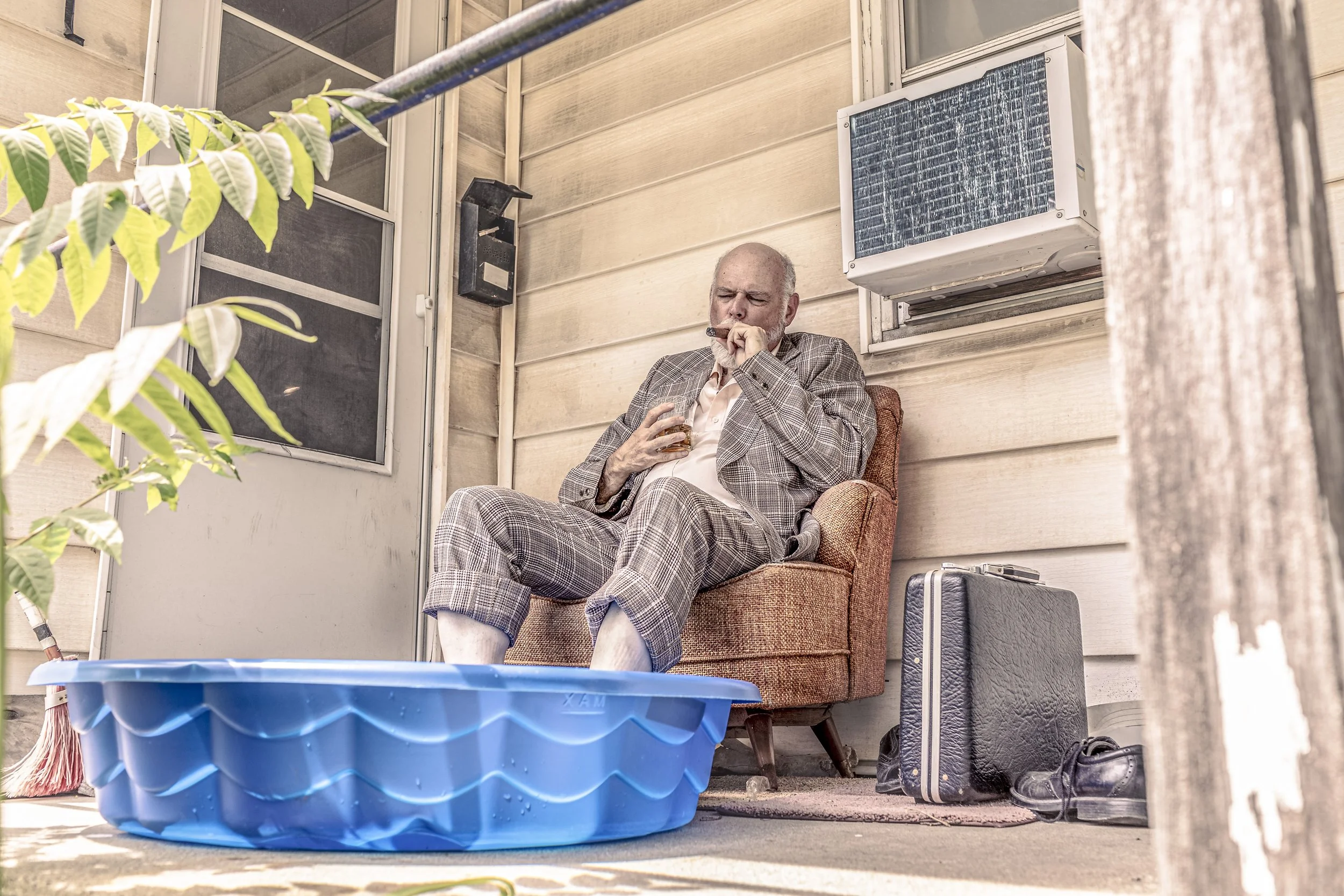 A  bald masc person in a rumpled suit sits in a chair on the porch with their feet in a blue, plastic swimming pool. Their eyes are squinted, and they’re puffing on a cigar and holding a glass tumbler full of an amber liquid.