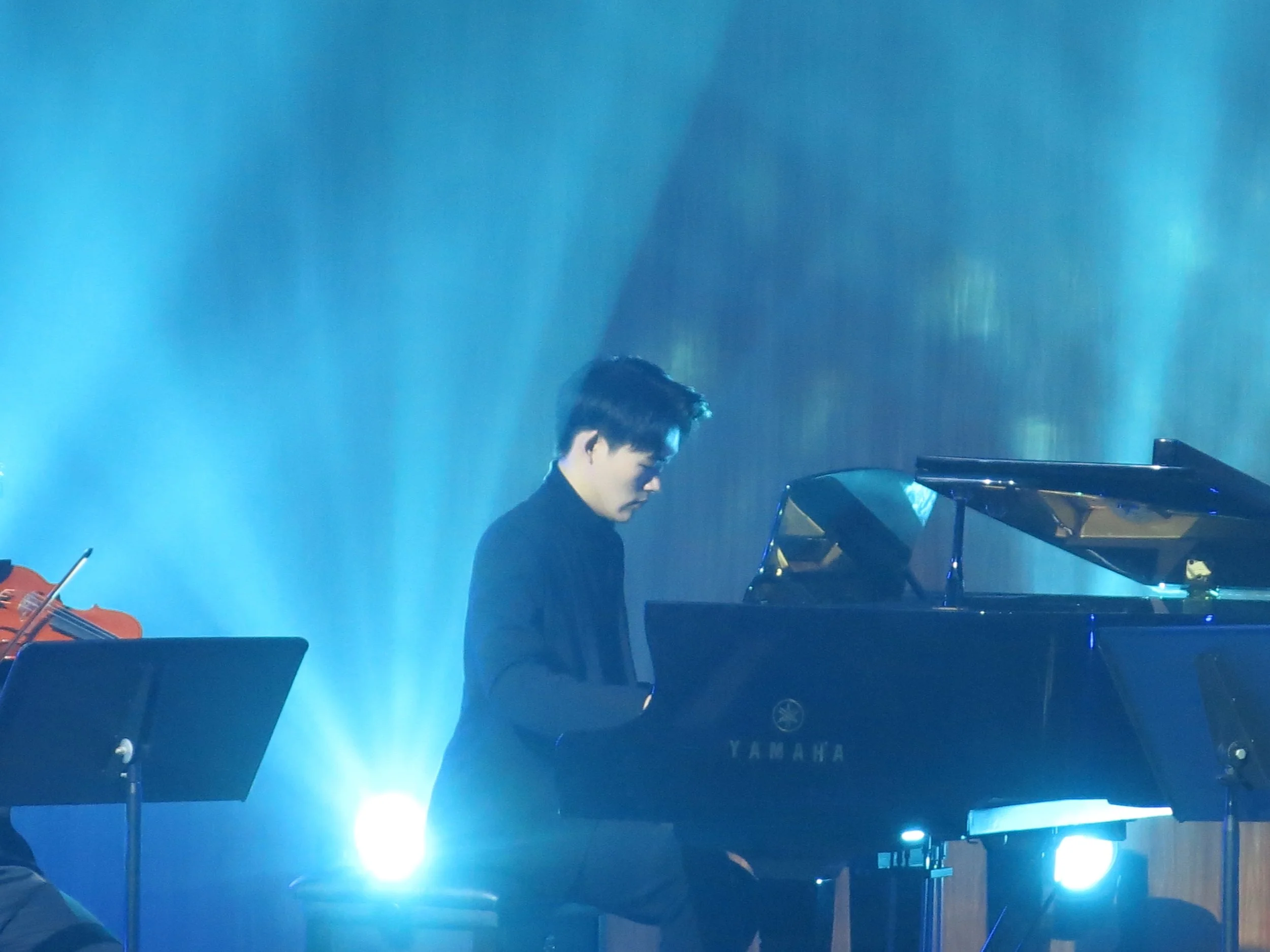 A person playing a Yamaha grand piano on a stage, illuminated by blue stage lights, with music stands and sheet music nearby.