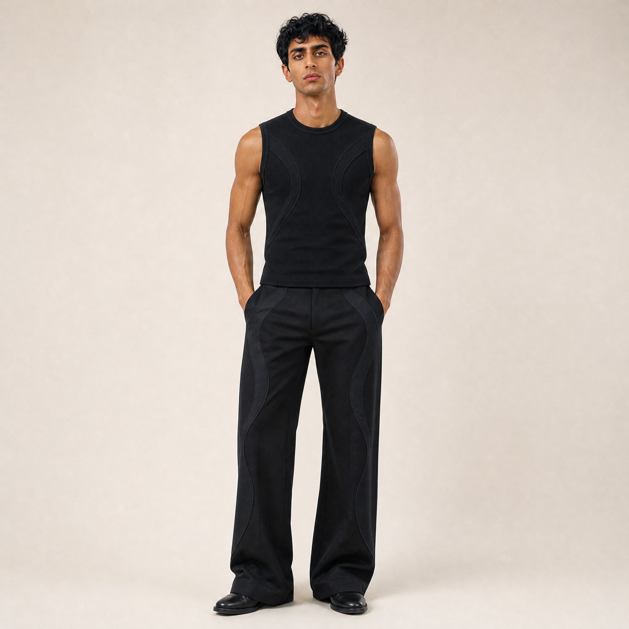 A tall man with dark curly hair and tan skin standing against a plain background wearing a sleeveless black top, black wide-leg pants, and black boots.