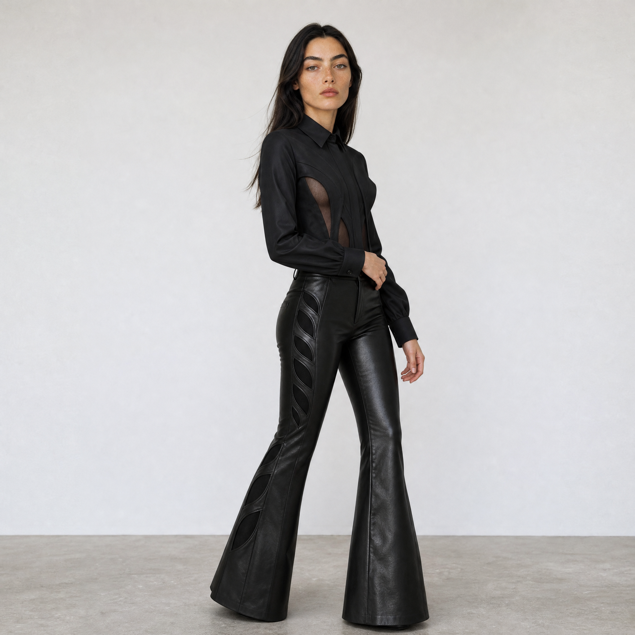 A woman with dark hair and fair skin, wearing a black long-sleeve shirt with mesh panels and black leather pants, standing against a plain light background.