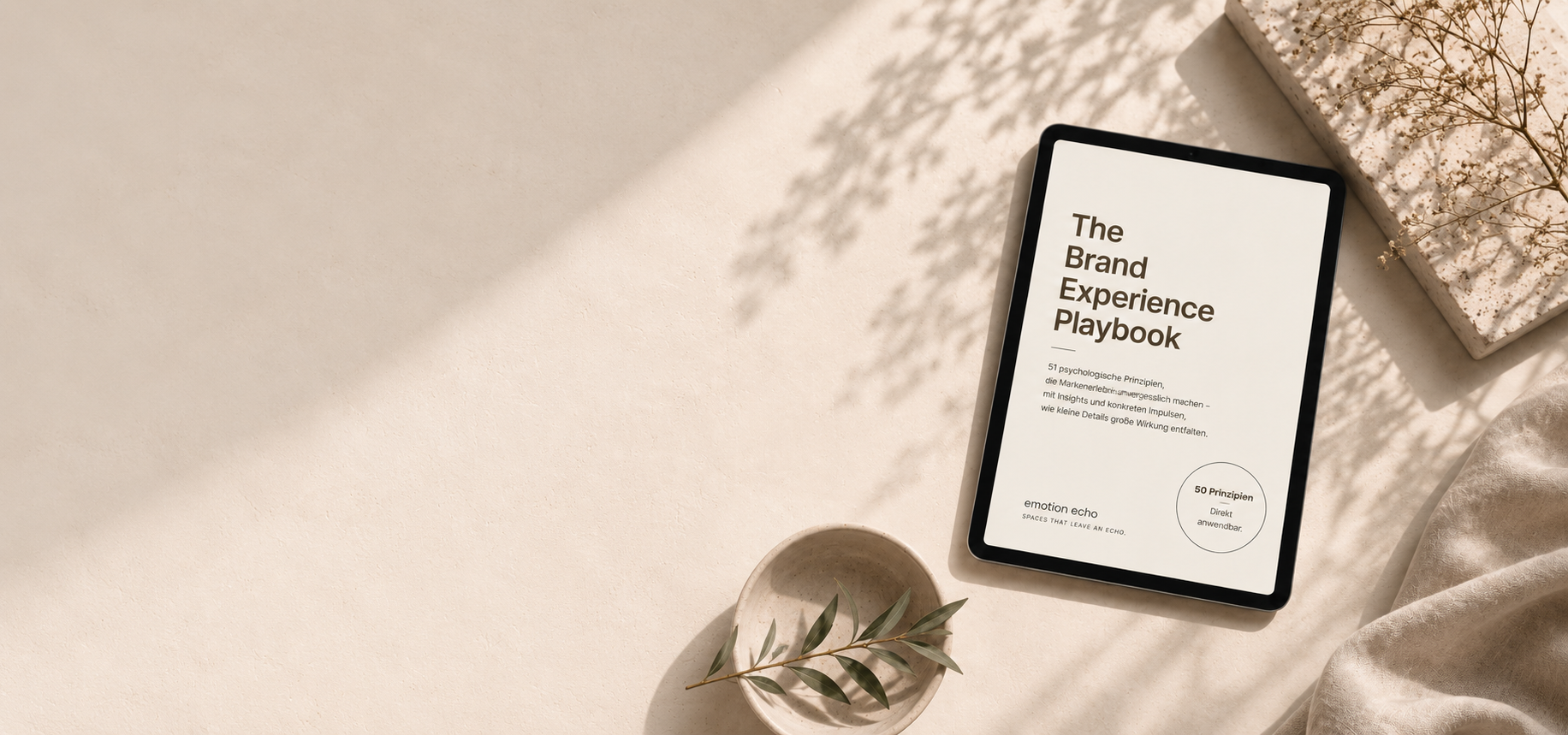 Ein Tablet zeigt das Cover eines Buches namens 'The Brand Experience Playbook' auf einem hellen Tisch, umgeben von einem kleinen Gefäß mit grünem Zweig, einem weißen Stein mit getrockneten Blumen und einem weißen Tuch.