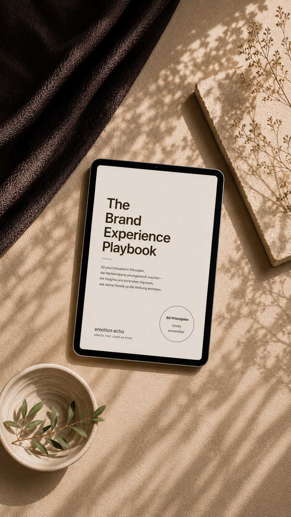 Ein Tablet zeigt den Titel 'The Brand Experience Playbook' auf einem beige Tisch. Daneben befindet sich eine Schale mit grünen Zweigen, im Hintergrund sind dunkler Stoff und eine weiße Block mit getrockneten Blumen. Schatten von Zweigen fallen auf die Szene.