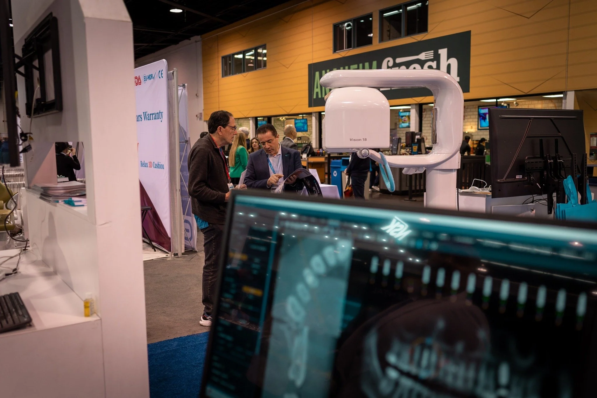 Two men talking at a technology exhibit booth with medical equipment and screens, in a large indoor convention center.