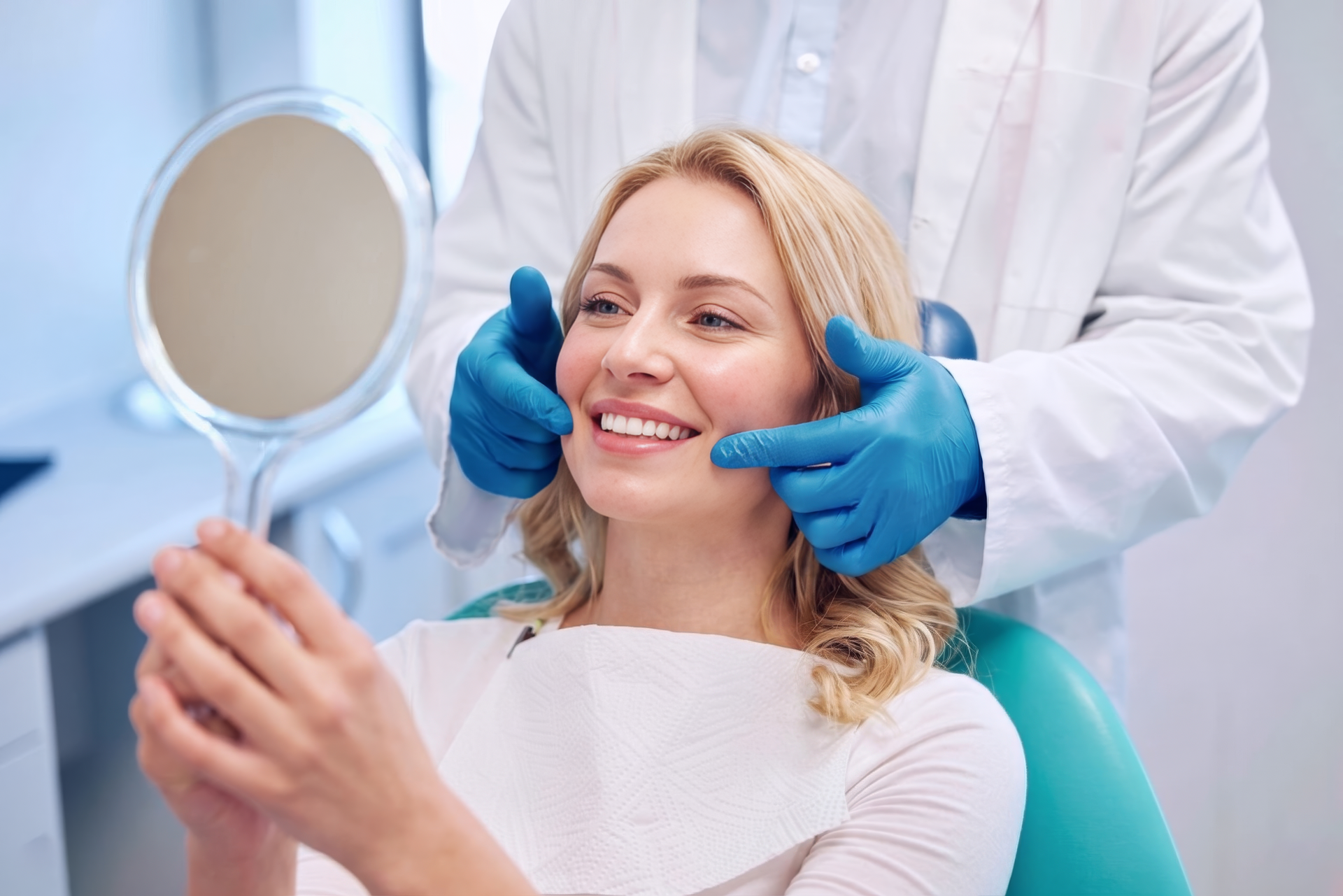 A woman sitting in a dental chair looking at a mirror, smiling. A dental professional in a white coat and blue gloves is gently holding her face.