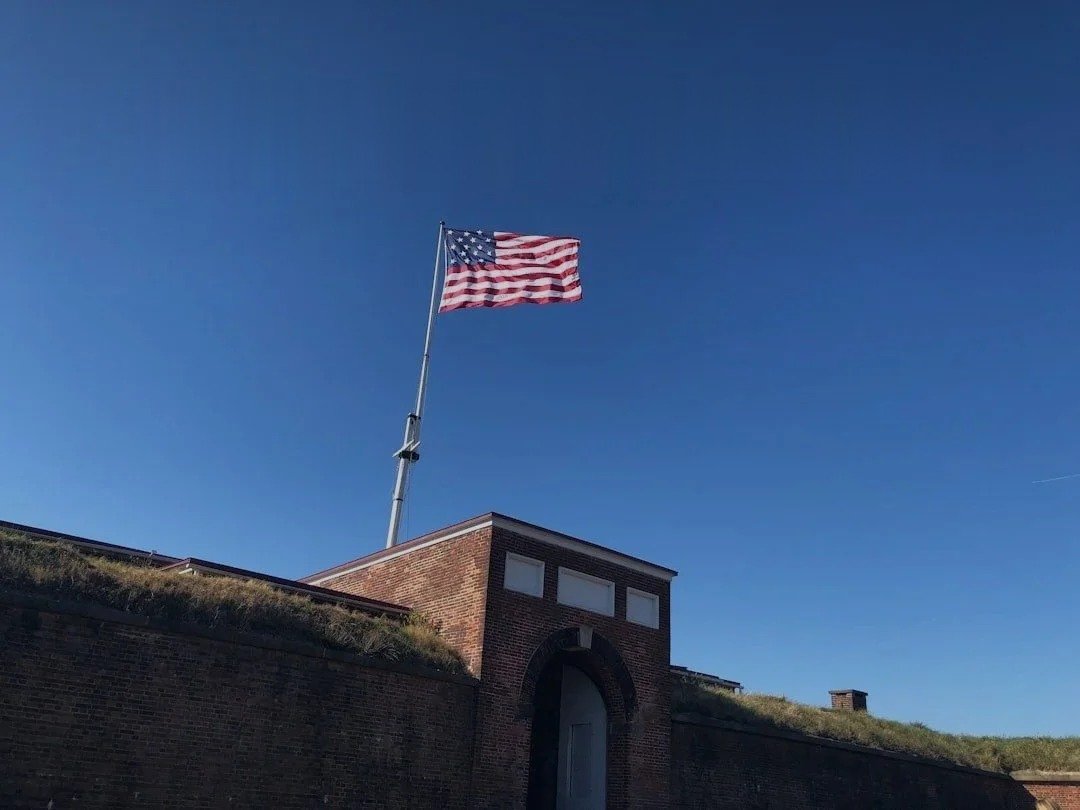 American flag flying on a flagpole above a brick fortress wall against a clear blue sky.
