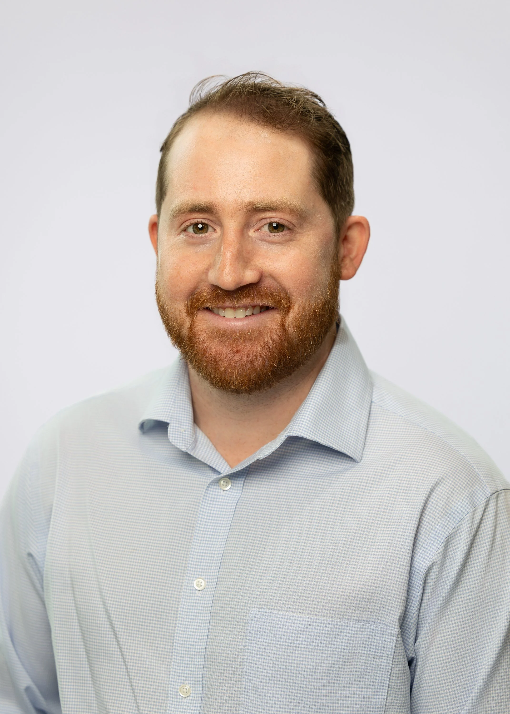 Headshot of a man with a beard and short brown hair, wearing a light blue dress shirt, smiling, against a plain light background.