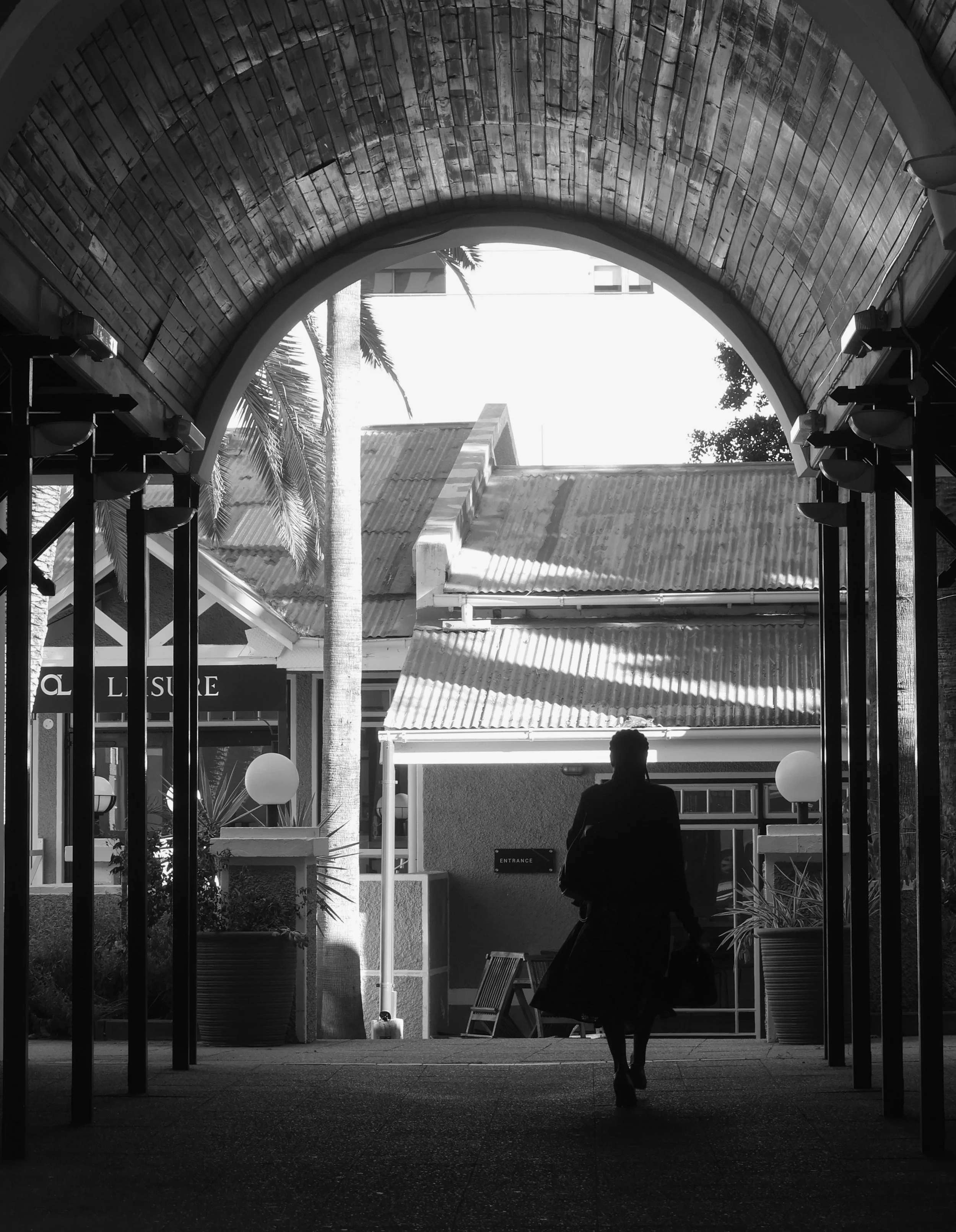 A black and white photo of a woman walking through an arched entranceway in a building, with sunlight outside illuminating palm trees and rooftops in the background. The woman is carrying a bag and is walking away from the camera.
