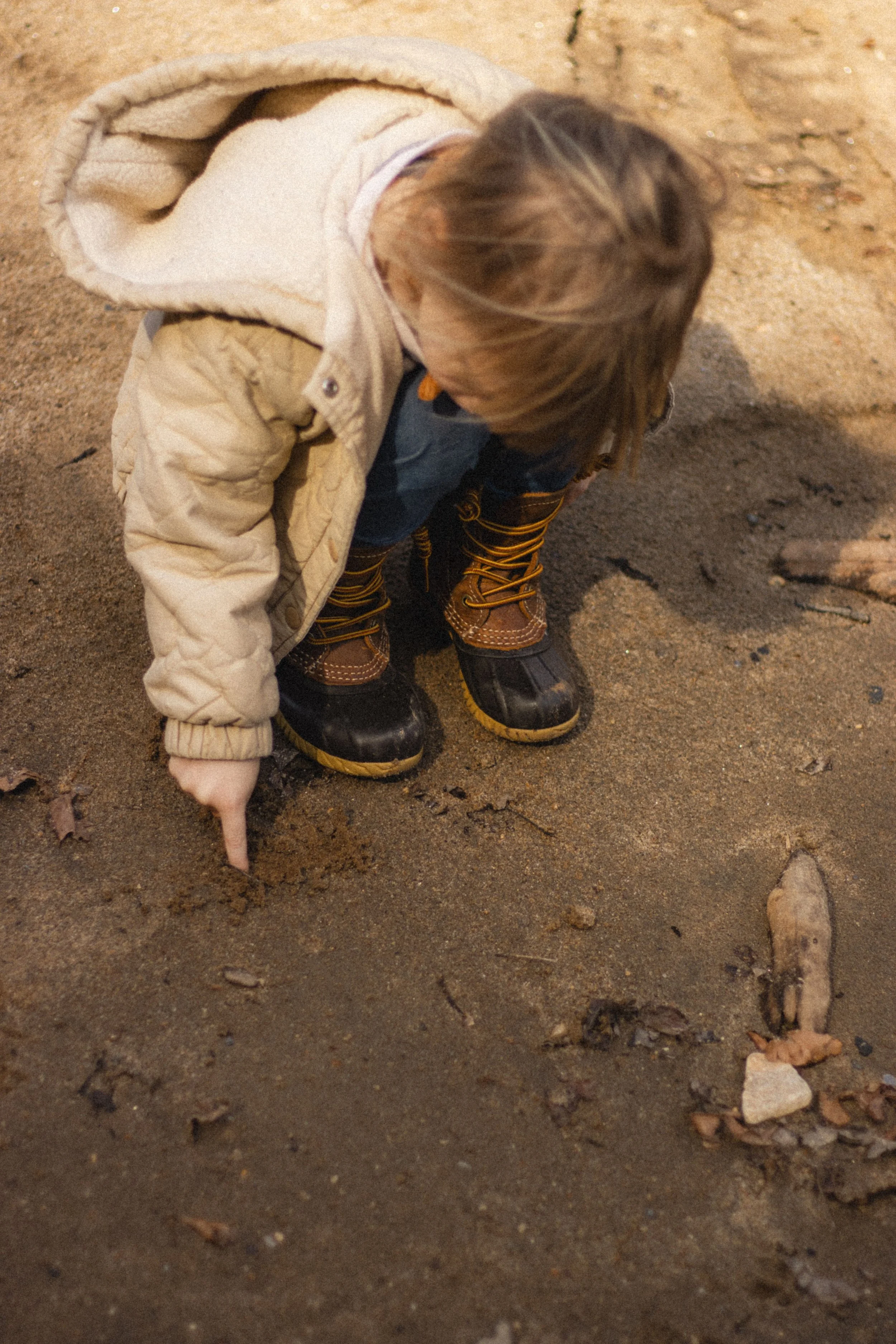 Young child with brown hair dressed in winter clothing, including boots and a beige jacket, holding a small white animal, possibly a rabbit, on sandy ground.