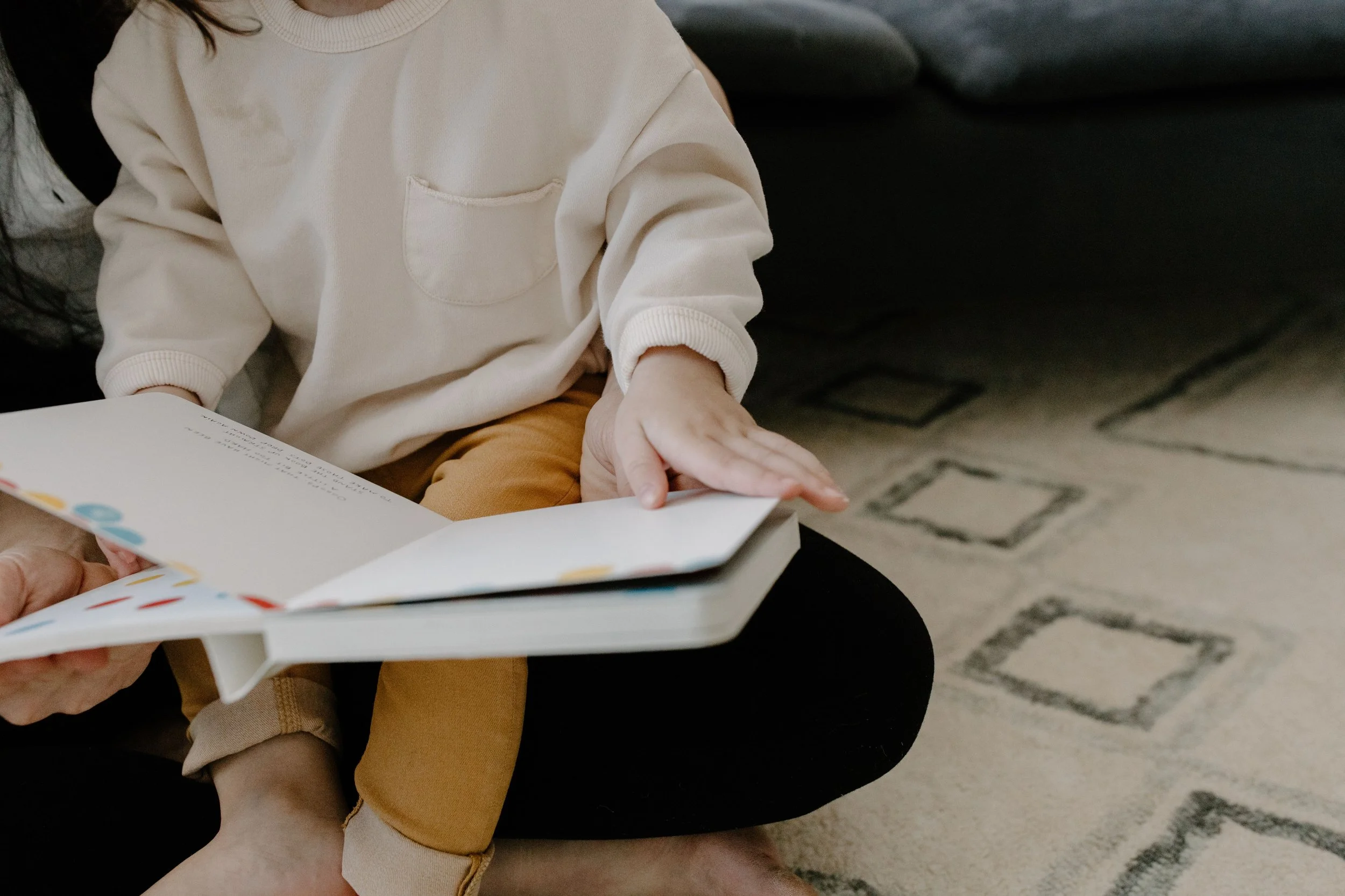 A person sitting on a couch, wearing a cream-colored sweatshirt and brown pants, holding and looking at an open book. Another person’s hand is holding the book from below.