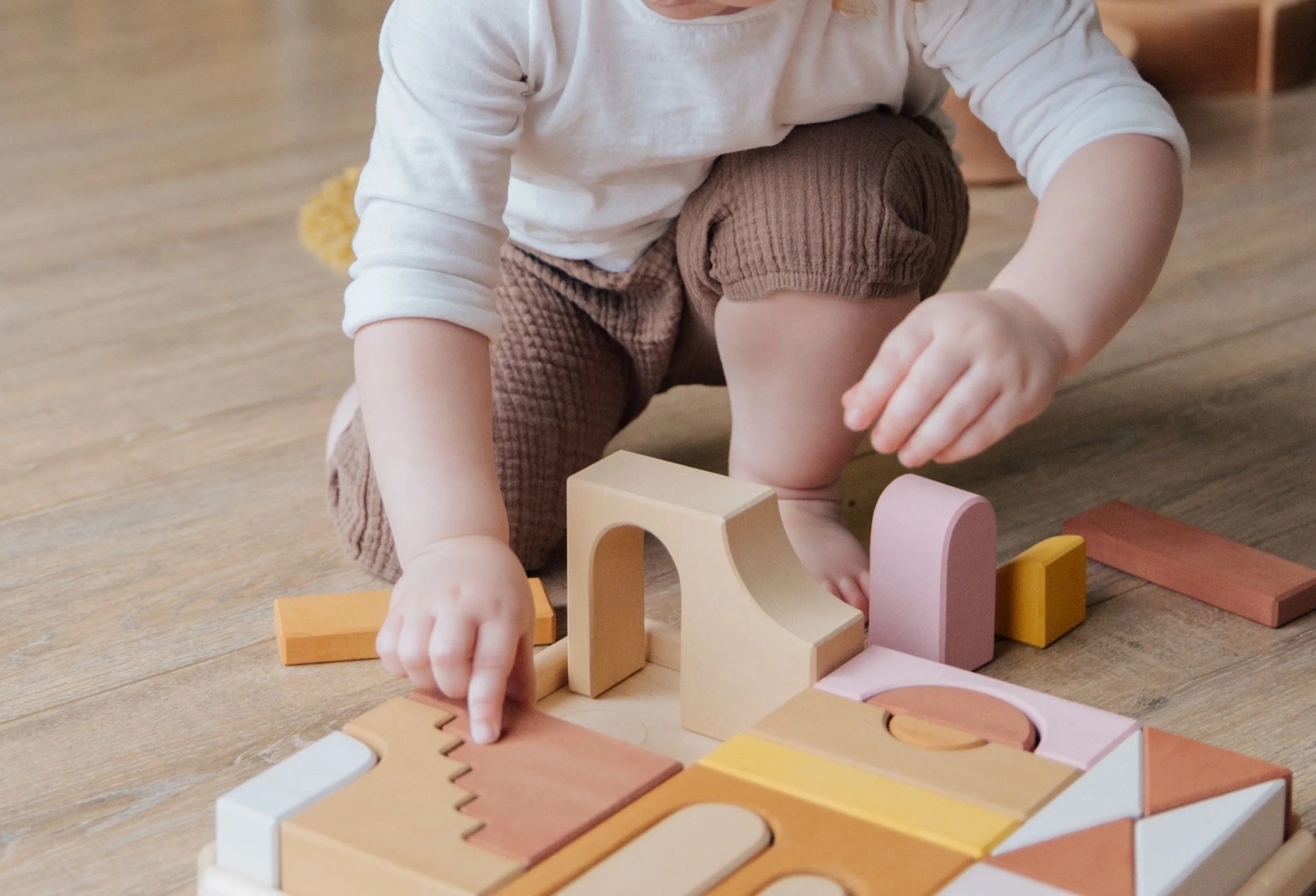 A young child dressed in a white long sleeve shirt and brown shorts playing with colorful wooden blocks on a wooden floor.