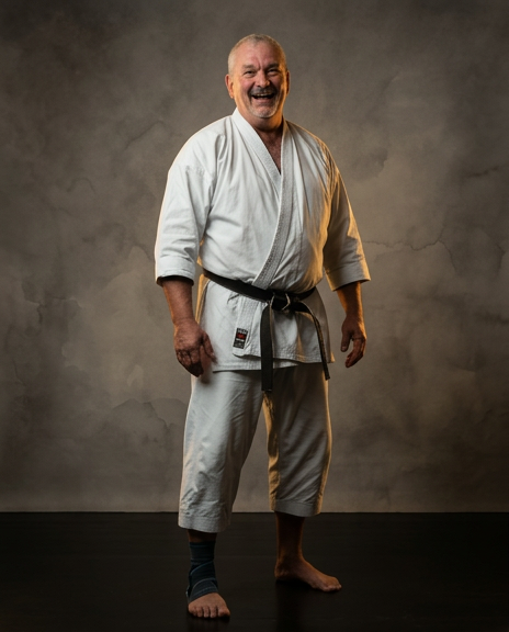 Un homme âgé en kimono de judo avec une ceinture noire, souriant, sur un fond gris.
