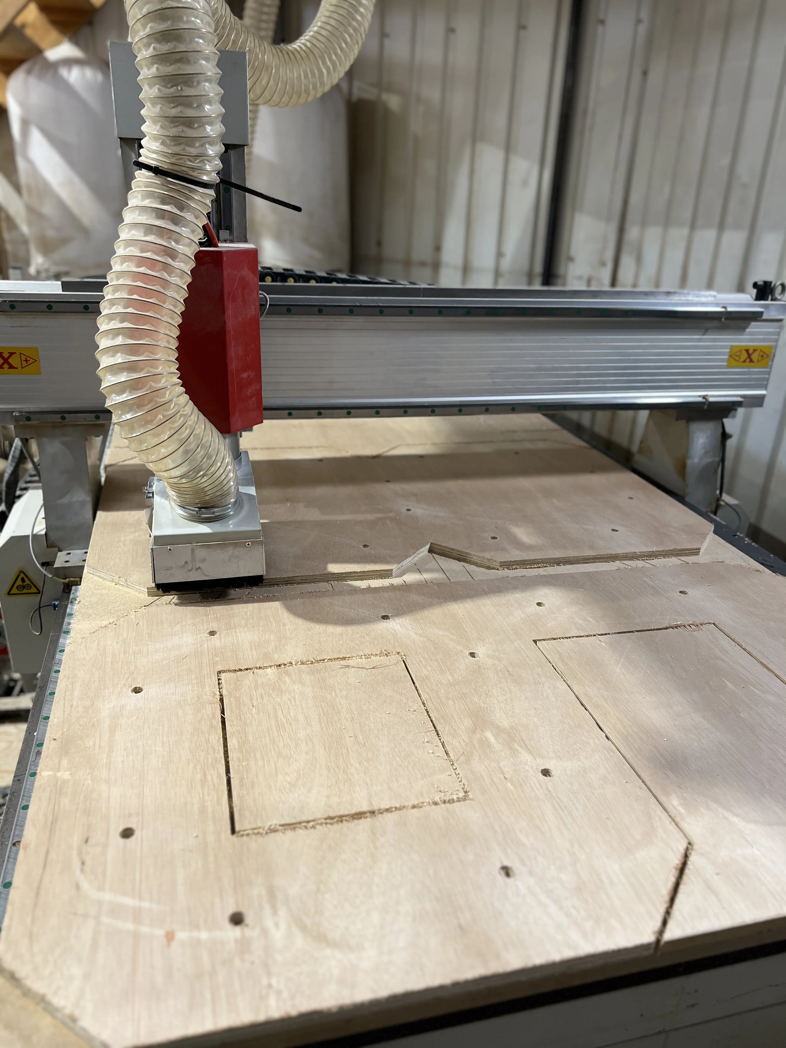 A woodworking CNC machine with a wooden workpiece on the bed, drilling or carving square shapes into the wood surface.