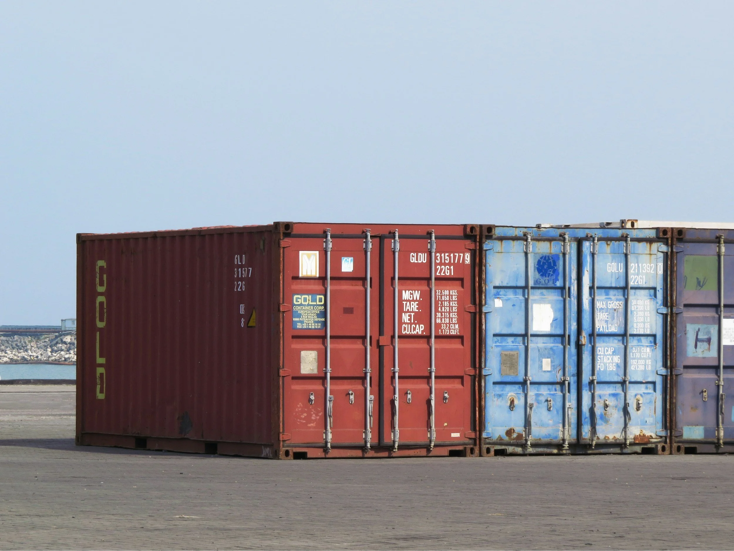 Three shipping containers, red and blue, sit side by side on a concrete surface with an open area behind them. The sky is clear and blue.