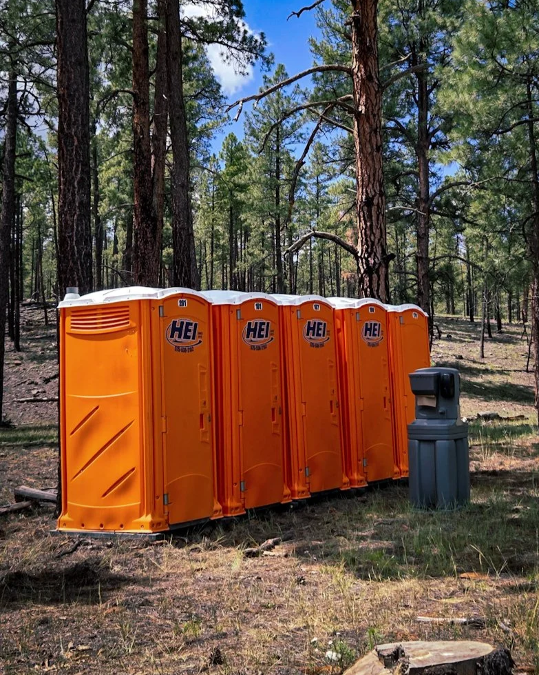 Four orange portable toilets with HEI logos are lined up in a forested area with tall trees and a partly cloudy sky.