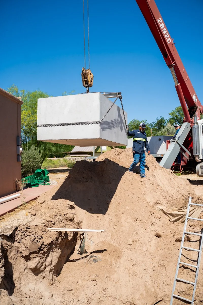 A construction worker standing on a pile of dirt next to a large concrete septic tank being lifted by a crane on a clear day.