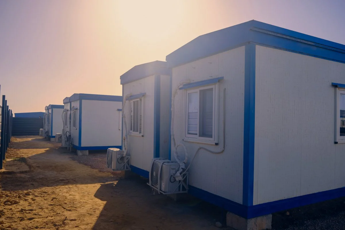 Multiple prefabricated white mobile offices with blue trim and air conditioning units, set in a sandy area with a strip of grass, under a clear sky at sunset.