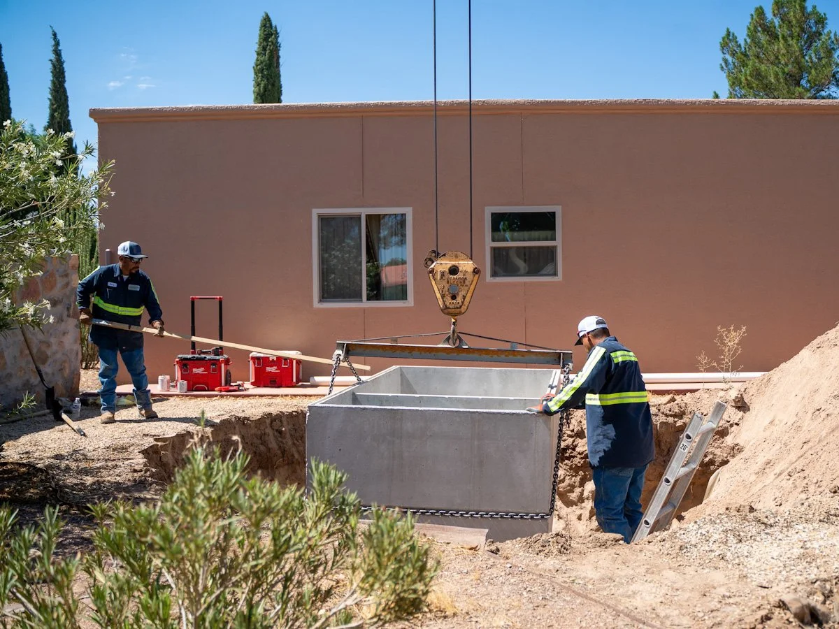 Two construction workers install a large concrete water tank in a backyard, with a house, trees, and a ladder in the background.