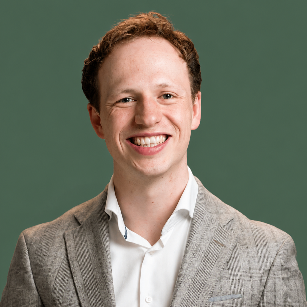 A smiling young man with red hair, wearing a gray blazer and white shirt, portrait against a green background.