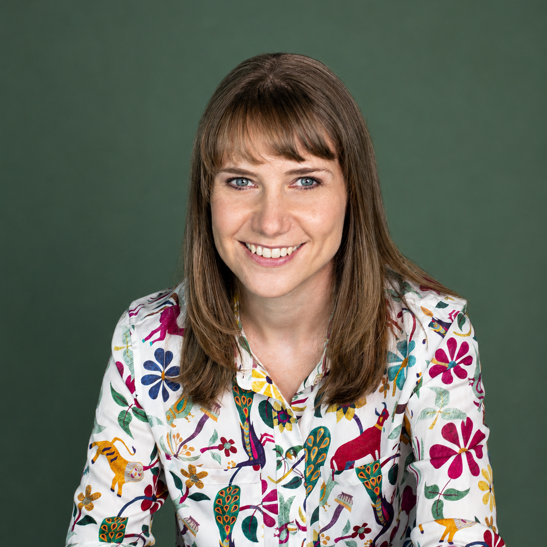 A smiling woman with straight light brown hair and bangs, wearing a colorful, patterned shirt with animals and flowers, against a blurred green background.