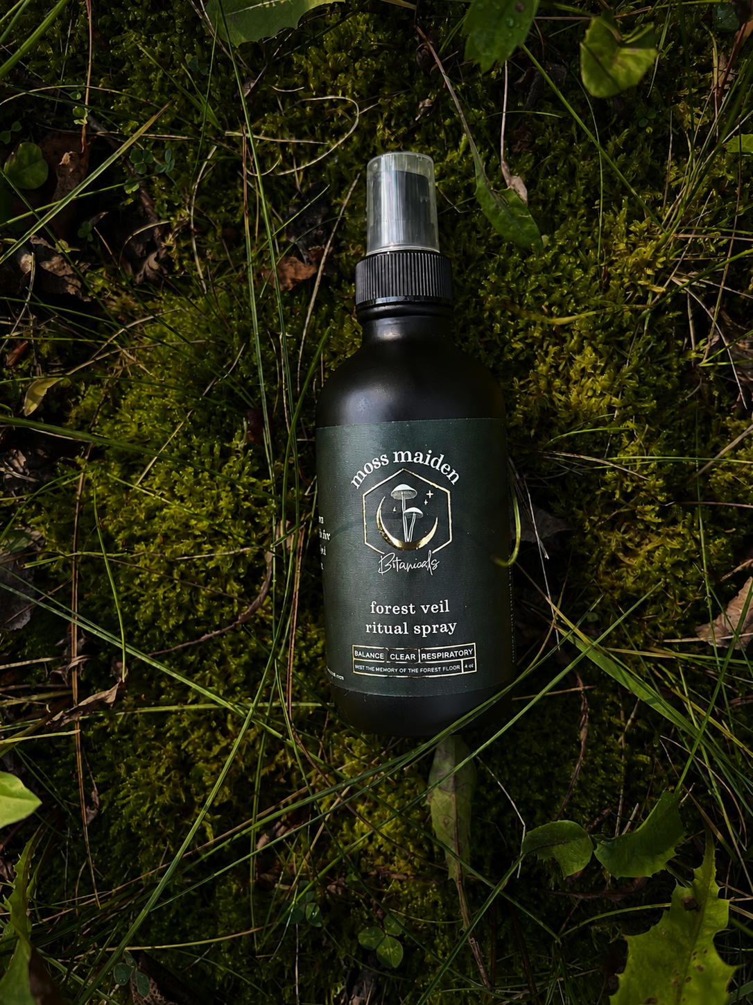 A bottle of moss maiden forest veil ritual spray lies on moss and grass in a natural outdoor setting.
