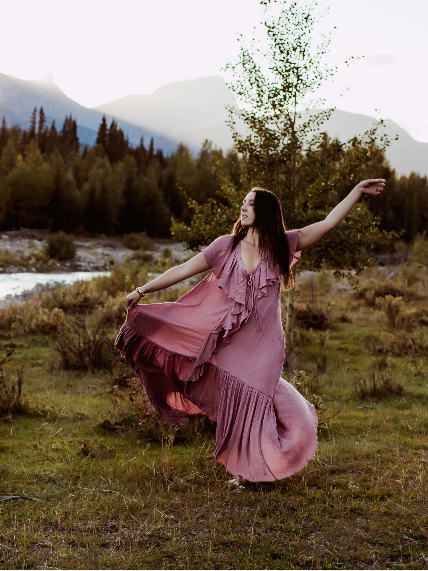 A woman in a flowing pink dress dancing or twirling outdoors near a grassy area with trees, mountains, and a river in the background during daylight.
