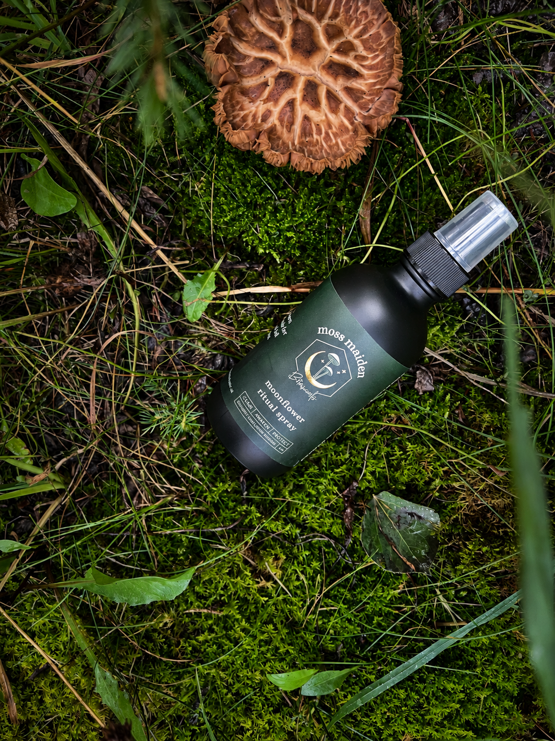 A black spray bottle labeled 'Moss Maiden' lying on moss and grass in a forest, with a brown mushroom nearby.