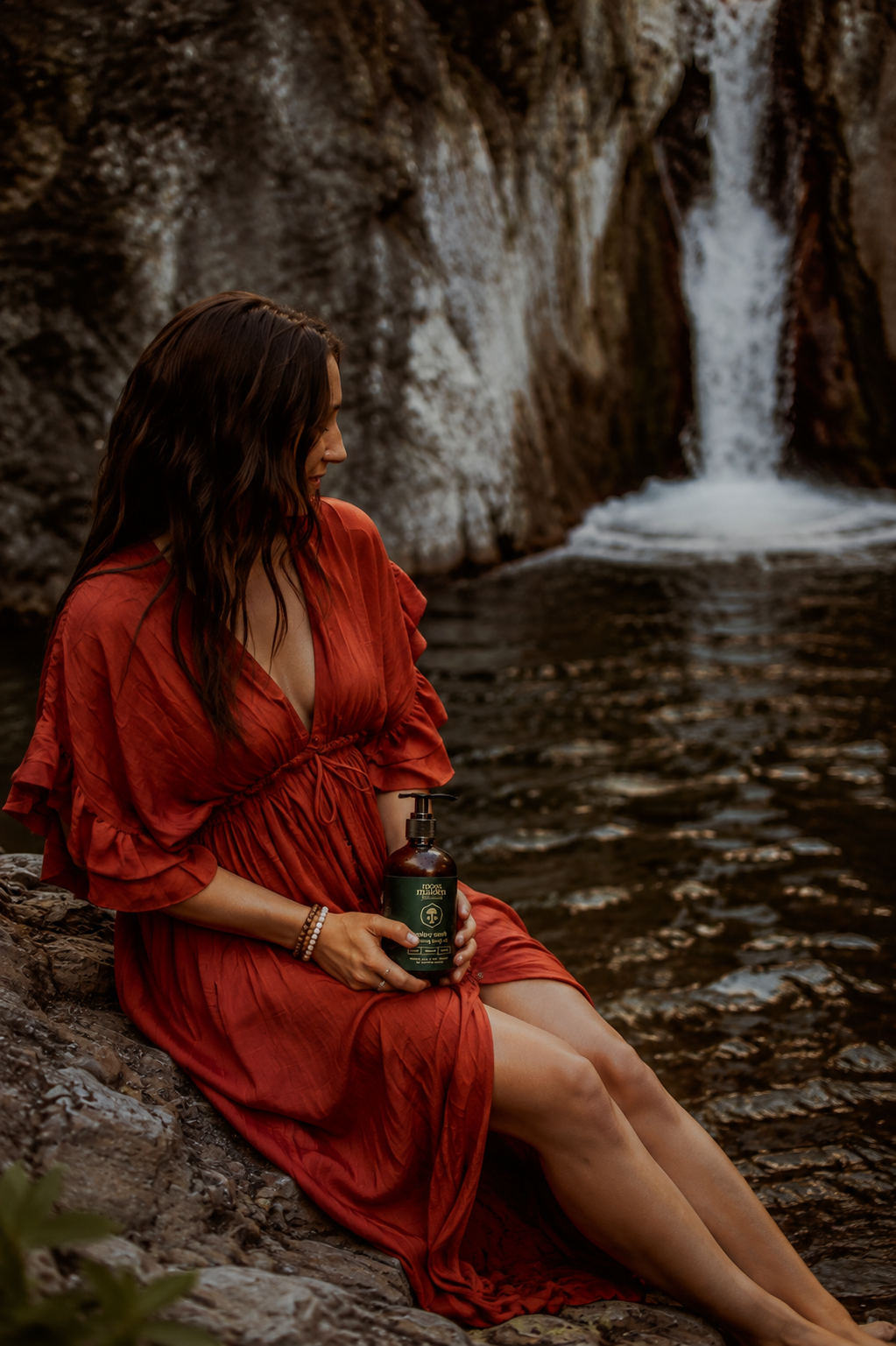 A woman in a red dress sitting on rocks by a waterfall, holding a bottle of hand soap.