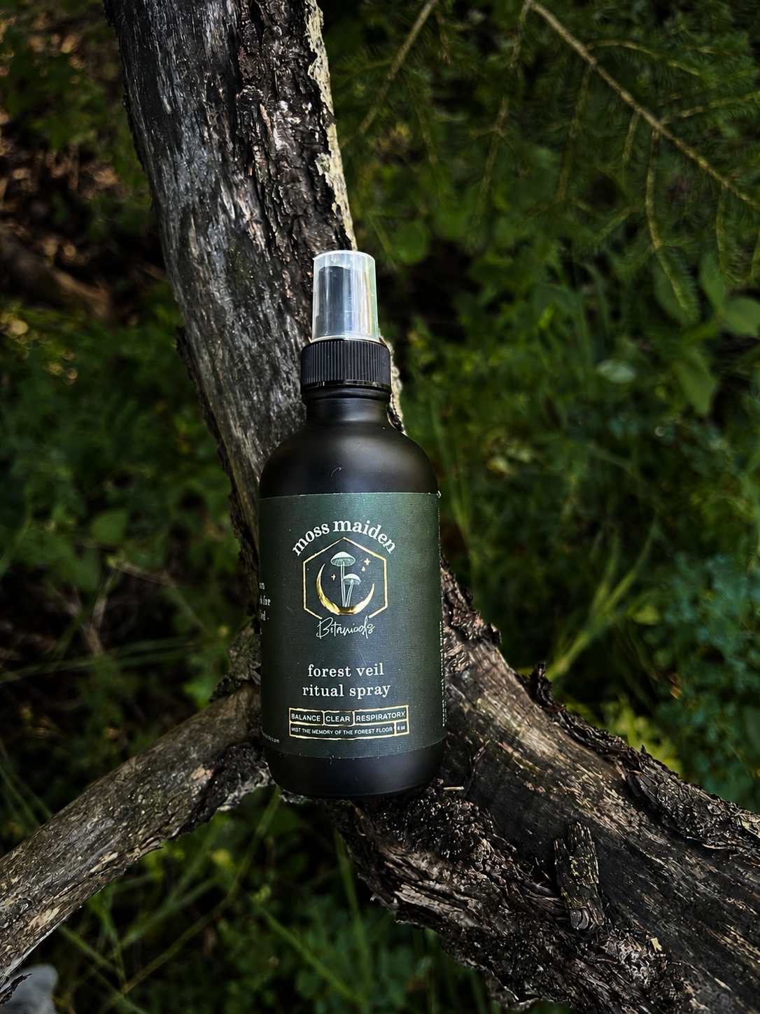 A spray bottle labeled 'Moss Maiden Forest Veil Ritual Spray' resting on a fallen tree branch in a forest setting.