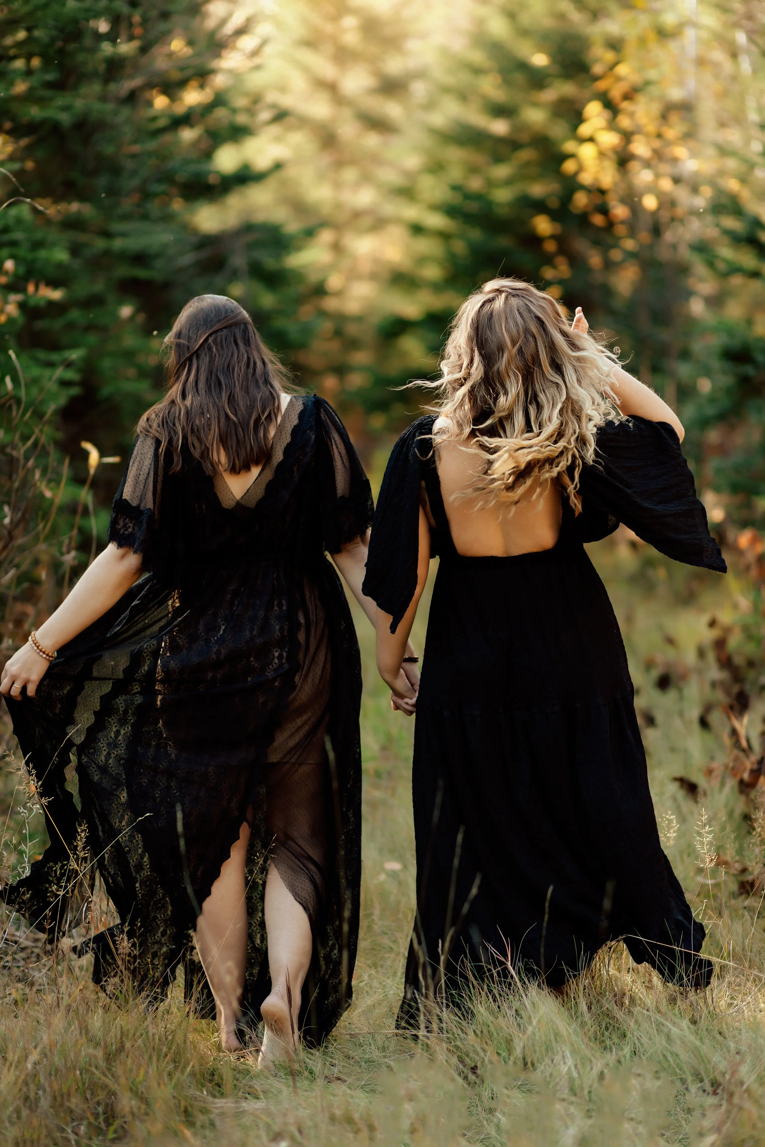Two women wearing long black dresses holding hands and walking through a grassy field with trees in the background during fall.